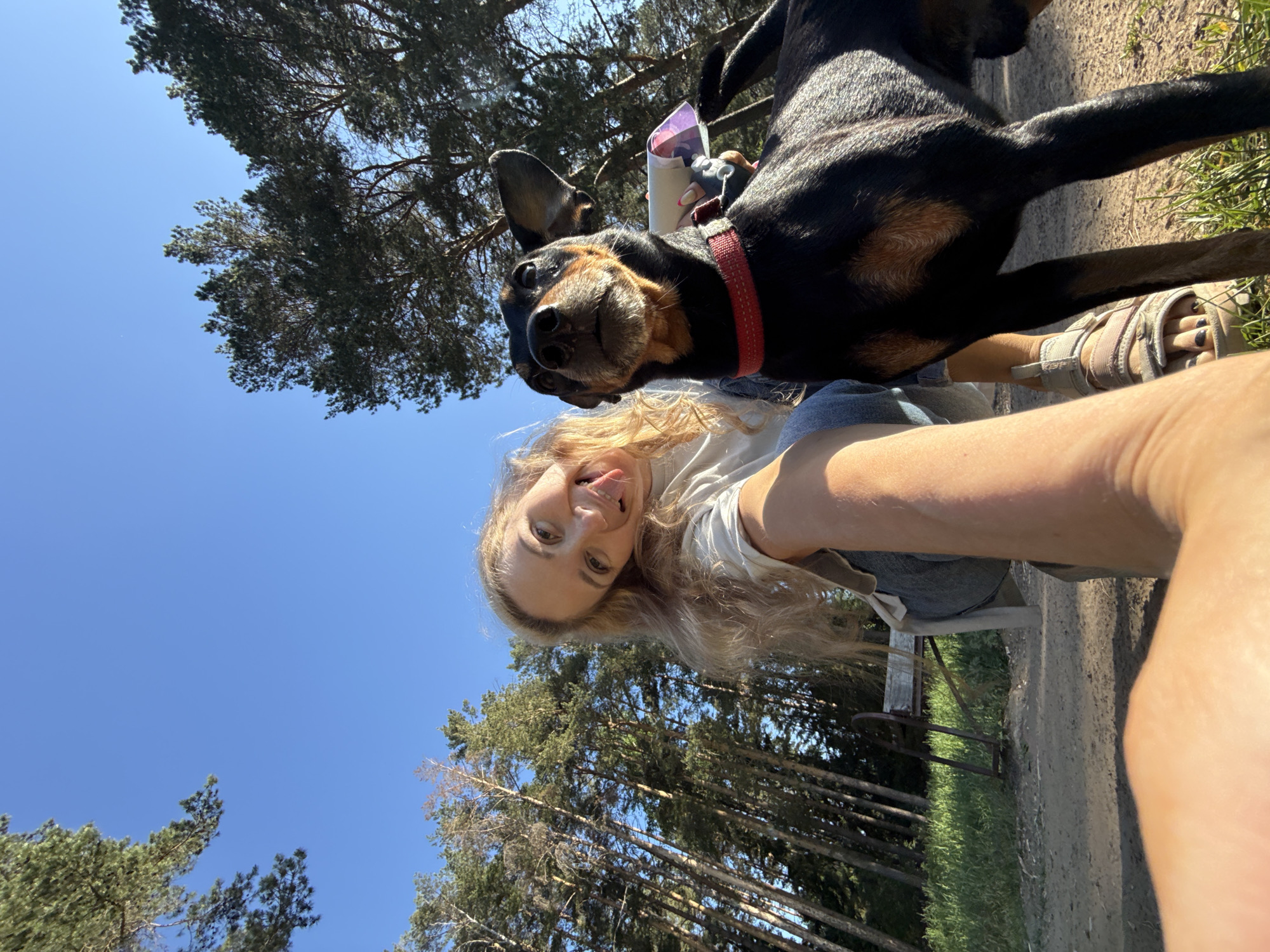 A selfie taken outdoors, likely in a park or wooded area. The subject, a young woman with long blonde hair, is holding the camera and sticking her tongue out. A black and brown dog with a red collar is next to her, looking into the camera. The sky is blue, with trees visible in the background. The location is the city of Chaikovsky, Russia.