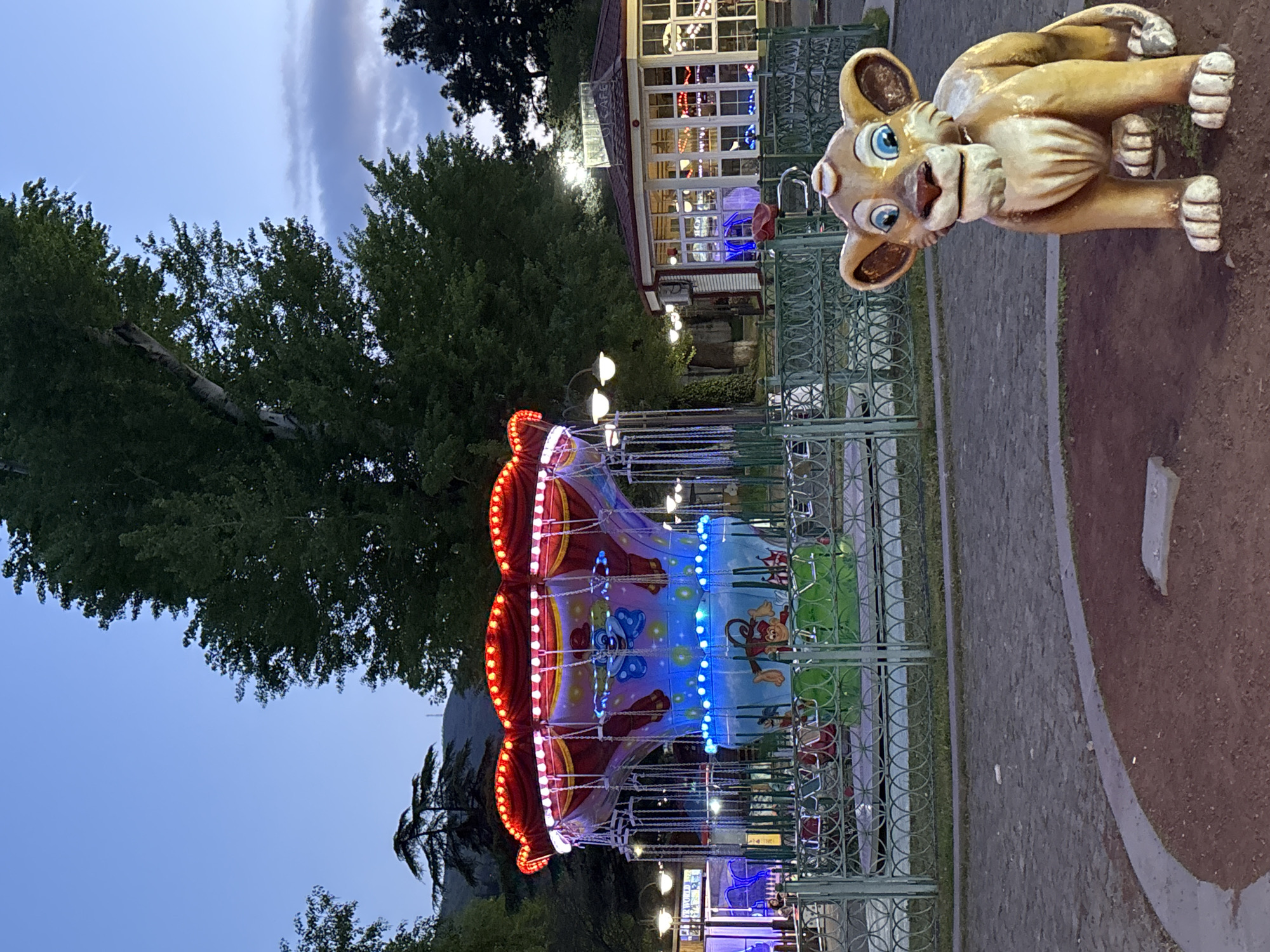The image depicts an illuminated amusement park or fairground scene at dusk or early evening. The sky, visible at the top and bottom, shows a transition from light blue to a darker twilight hue, with some faint cloud streaks.
A prominent feature is a children's swing carousel ride, centrally located, which is brightly lit with numerous red and blue LED lights. The ride's canopy and panels are decorated with colorful illustrations of cartoon characters, including a monkey and a butterfly. The swings of the carousel appear empty.
To the right, a large, light brown, cartoon-style statue of a lion or similar feline rests on the ground. The statue has distinctive blue eyes and a smiling or friendly expression. It lies beside a paved pathway.
In the background, a building with many windows, possibly a cafe or restaurant, is visible, brightly lit from within. A sign above its entrance appears to read "CAFE". Lush green trees frame the scene, creating a natural park-like environment. Ornate green metal fences delineate sections of the park and pathways. The ground consists of dark soil and paved sections. The scene conveys a lively, recreational atmosphere in Tbilisi, Georgia, as artificial lights begin to dominate with the setting sun.