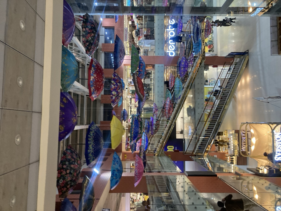 This image depicts the interior of a multi-story indoor shopping mall or commercial complex in Quetta City Tehsil, Pakistan. The primary visual element is a dense overhead installation of numerous brightly colored, inverted umbrellas suspended from the high, grid-patterned ceiling throughout the central atrium. These umbrellas display a variety of patterns and solid colors, including blue, yellow, purple, red, and floral designs.
The facility features multiple levels, connected by prominent escalators with glass balustrades and metal handrails. Upper walkways are bordered by glass railings. The flooring is composed of polished light-colored tiles, and some structural columns and walls are finished in orange-brown or white.
Several individuals are present, observed in various actions of movement and presence within the mall. On the ground floor, a person wearing a dark jacket is visible near a kiosk displaying "SVESTON" and "MOXAZ" branding. To the right, an adult carries a small child, accompanied by another child walking alongside. Other individuals are visible on upper levels and utilizing the escalators. Retail establishments are discernible on different floors, including one with a large blue "den obe" sign and another partially legible sign indicating "ACHAKZAI CLOT." The environment is illuminated by recessed ceiling lights and interior shop lighting.