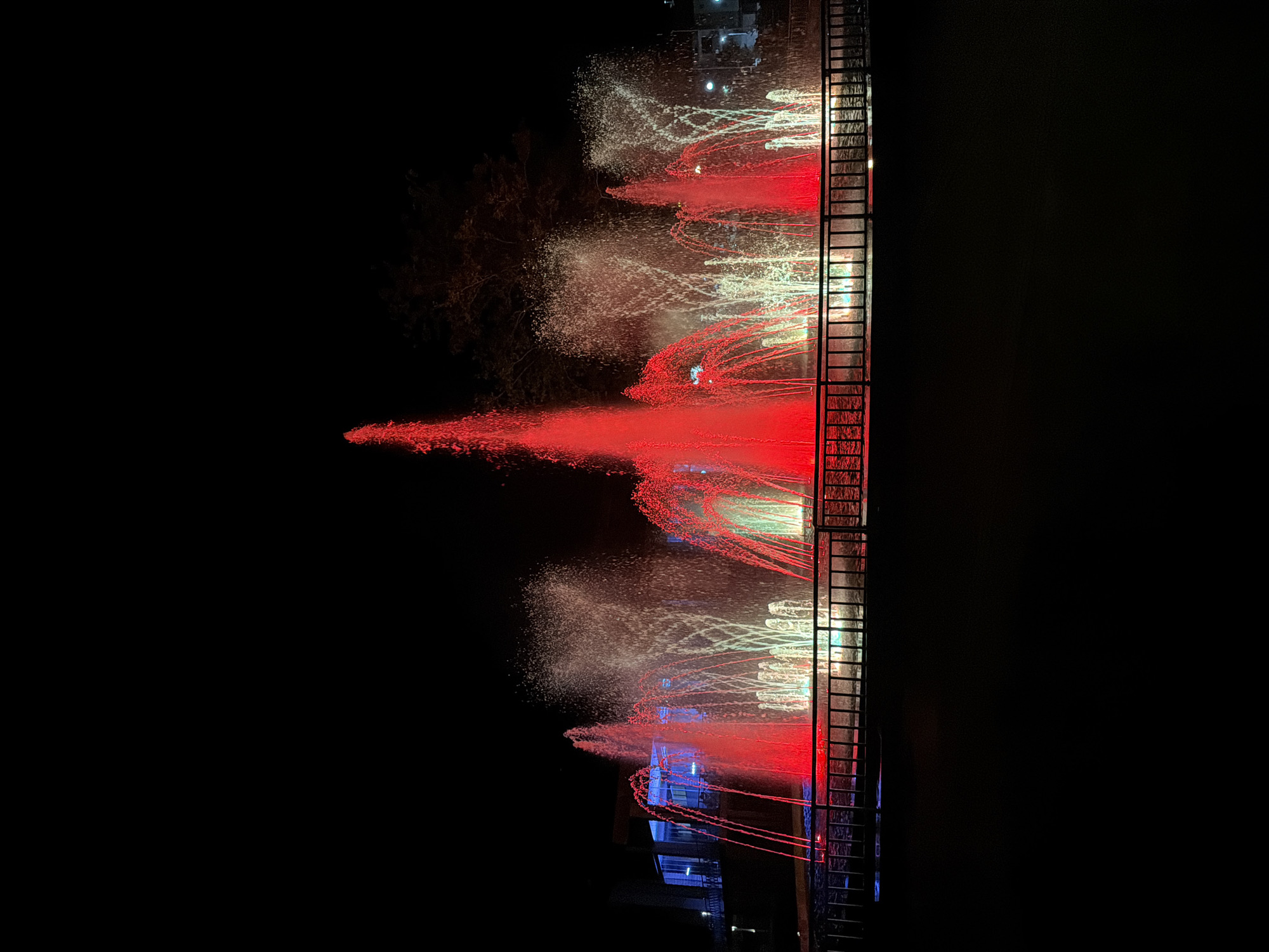 A nighttime view shows a fountain display in the city of Panagyurishte, Bulgaria. The fountains are illuminated in red and white. A ladder extends vertically along the right edge of the fountain display. The background is dark, with faint building lights visible at the top. A bare tree is partially visible on the left.