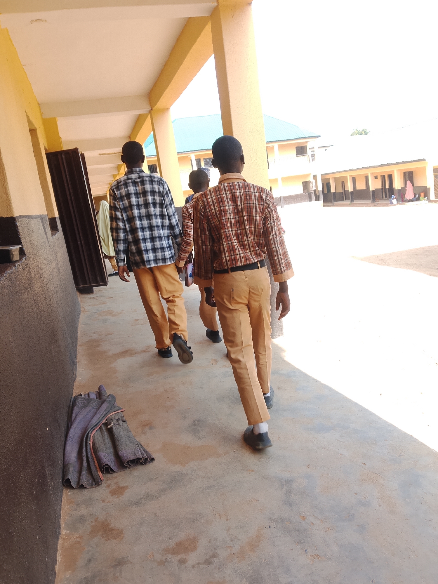 The image shows three school-aged individuals walking away from the camera down an outdoor covered walkway. The individuals are dressed in school uniforms consisting of plaid shirts and light brown pants. The walkway is paved and has yellow-painted pillars supporting a white ceiling. To the left of the walkway is a dark brown wall. A pile of dark grey and pink fabric is on the ground near the wall. In the background, across a courtyard, are several buildings with light yellow walls and dark brown trim. The sky is bright and appears to be a sunny day. The location is described as Unknown, Unknown.