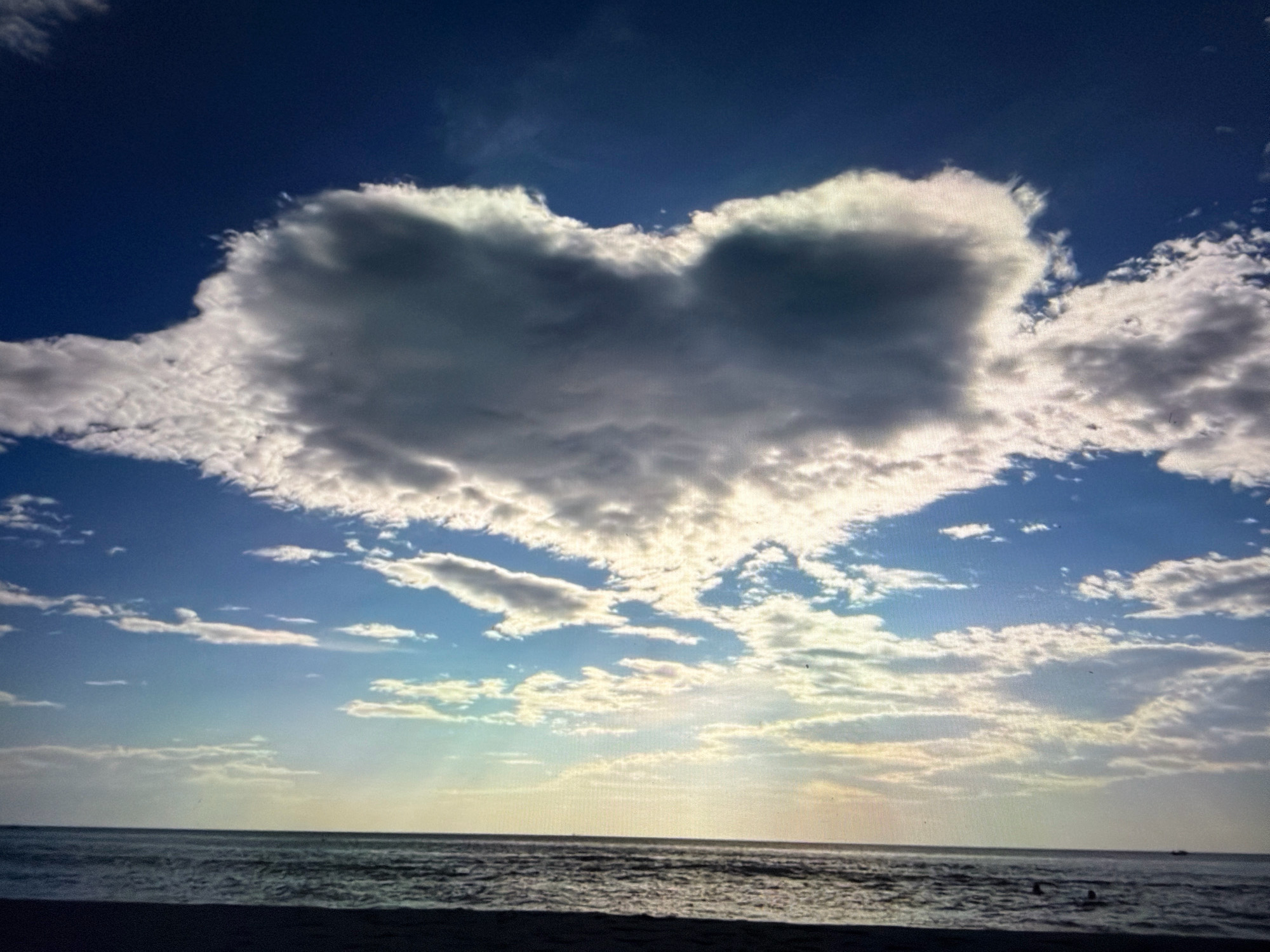 The image depicts a beach scene with a prominent heart-shaped cloud in a deep blue sky. The ocean is visible in the foreground with gentle waves, and a dark beach stretches across the bottom of the frame. The lighting suggests it could be sunrise or sunset, with the sun casting a glow on the horizon and illuminating some of the clouds. There are no specific landmarks or identifiable features that would pinpoint the location to Đà Nẵng, Vietnam. The image primarily focuses on the natural elements of the sky, sea, and a unique cloud formation.
