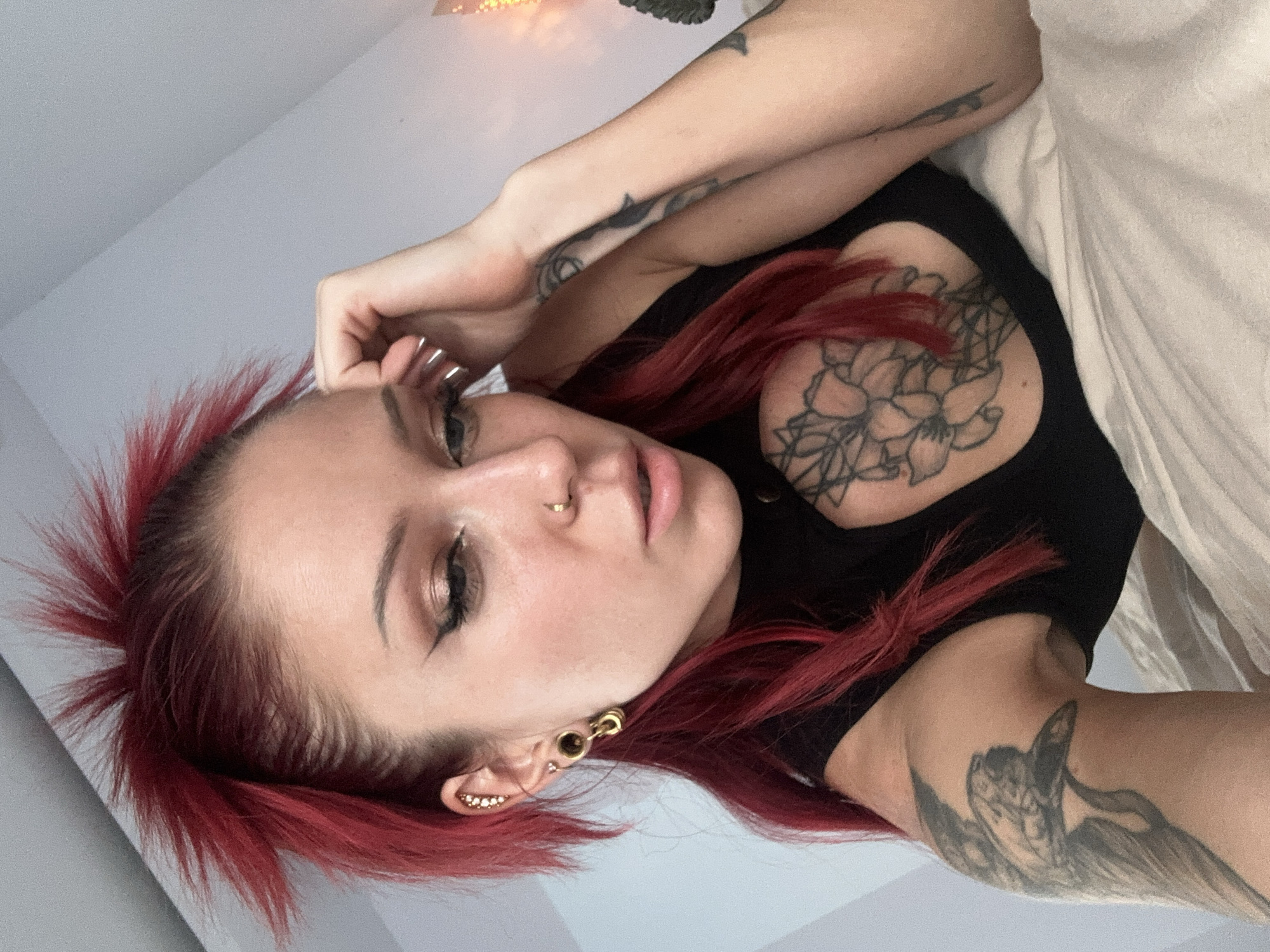 A woman with red hair lies on a bed. She has multiple tattoos on her arms and chest. She has a nose piercing and multiple ear piercings. She is wearing a black shirt. The background is a white wall. The location is not specified.