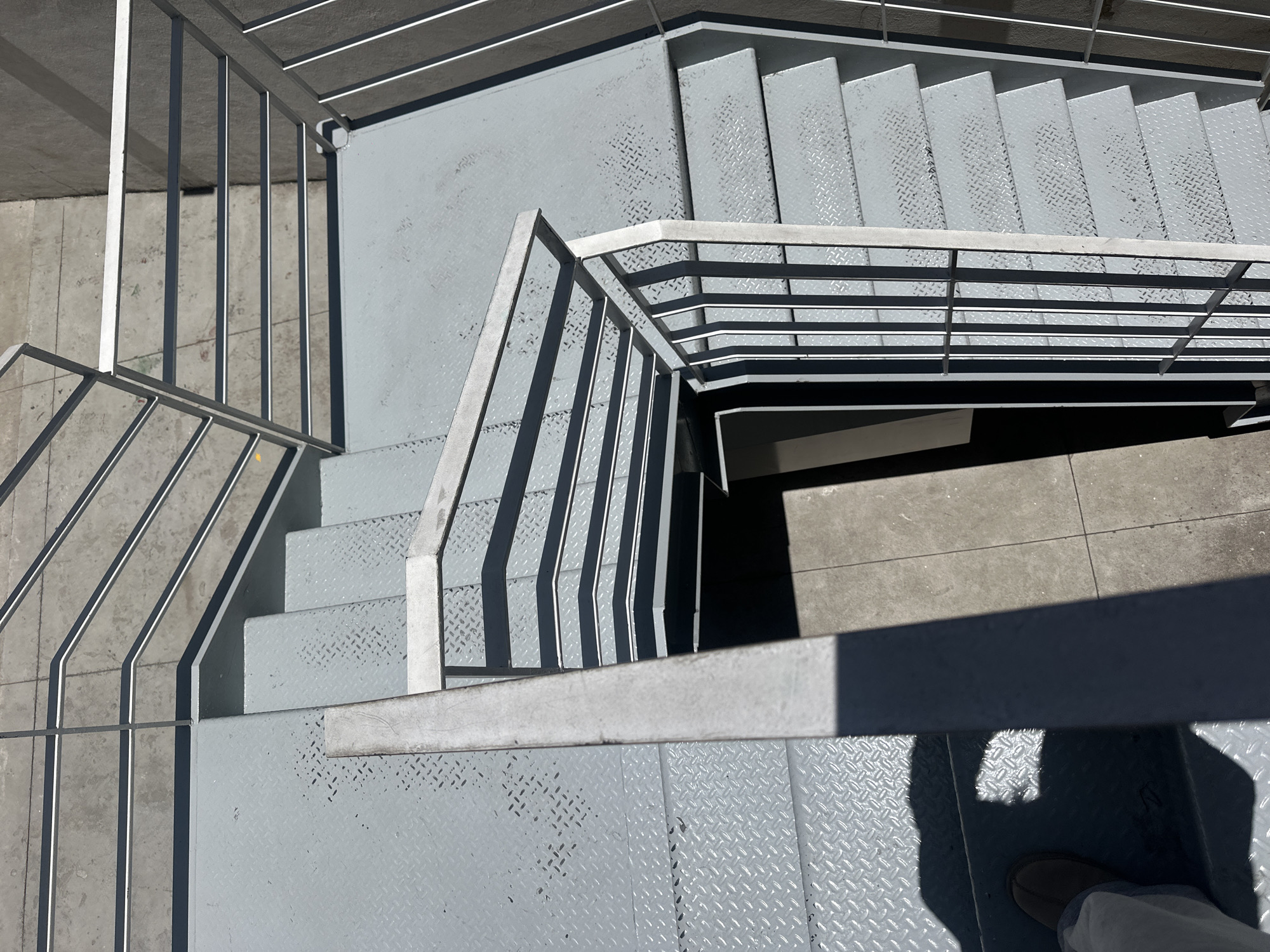 The image shows an elevated perspective of metal stairs with handrails. The stairs are painted gray and feature a diamond plate pattern on the treads and landing. The handrails are made of metal with vertical bars. A shadow of a person is visible in the lower right corner of the frame, suggesting the image was taken by someone standing on the stairs. The location is Mexico City, Mexico.
