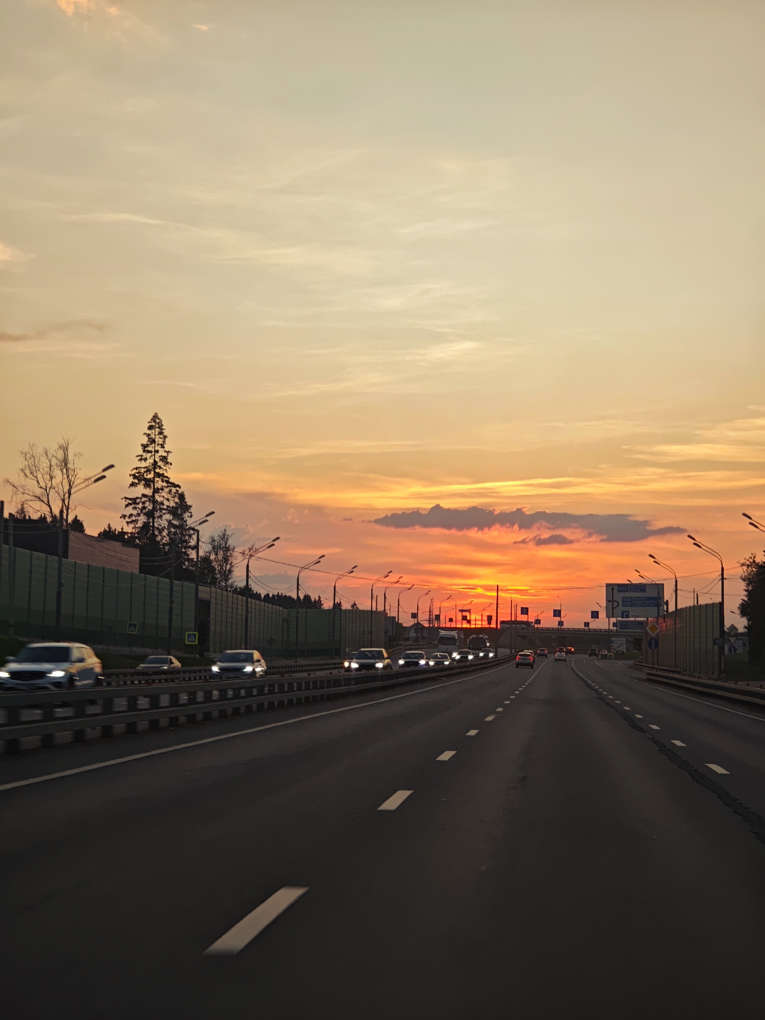 The image captures a highway scene at sunset, likely in or near посёлок опытного хозяйства «Ермолино», Russia. The sky is ablaze with vibrant orange and yellow hues, indicative of a dramatic sunset, with scattered clouds adding texture. A multi-lane highway stretches into the distance, bordered by sound barriers on the left and right. Numerous cars are visible on the highway, most in motion and appearing as streaks of light due to the long exposure or motion blur. Streetlights line the highway, and trees and foliage are visible beyond the sound barriers, suggesting a suburban or semi-rural setting. Traffic signs are present along the roadside. The perspective is from a vehicle moving along the highway.