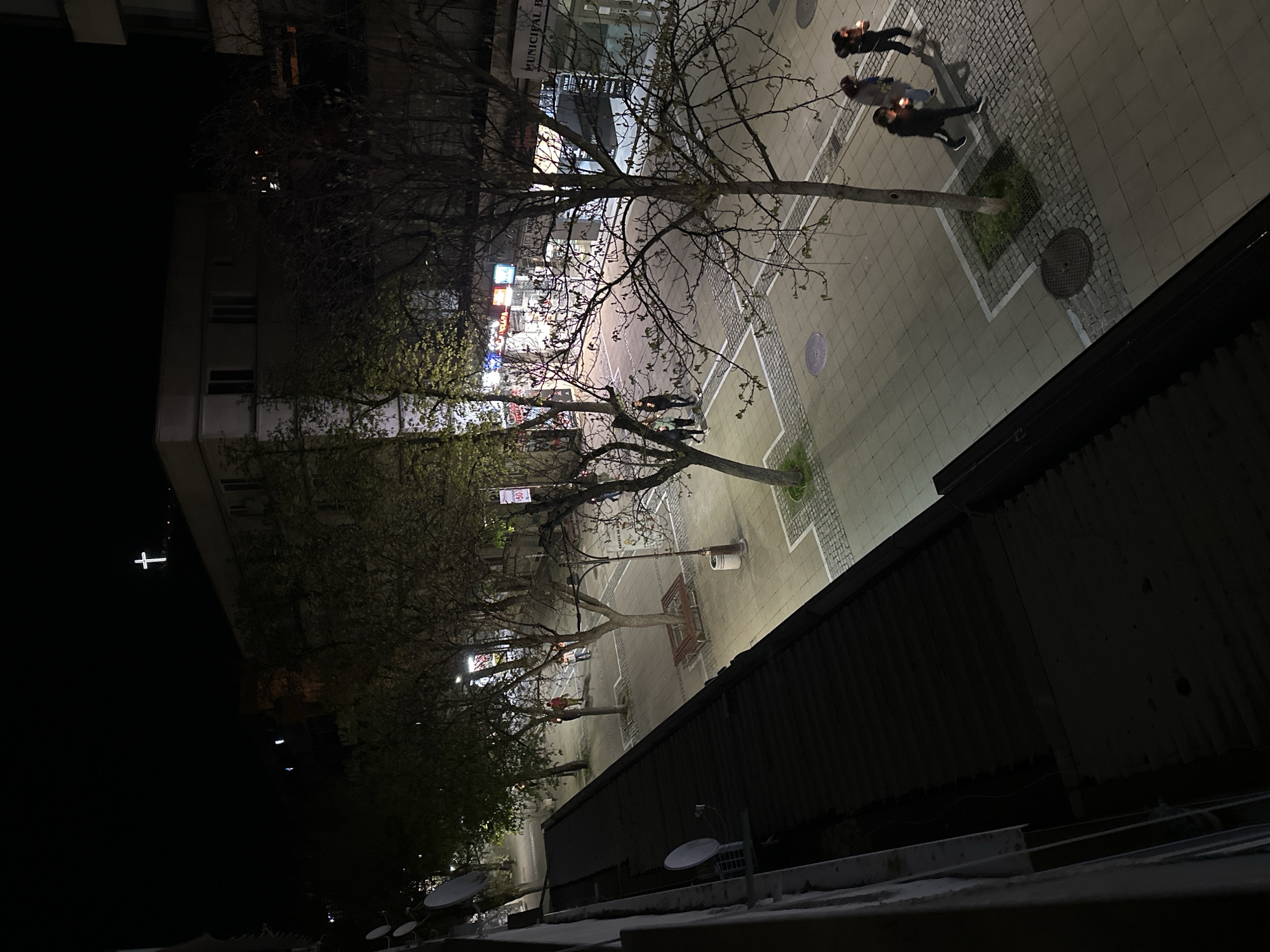 The image shows a nighttime view of a pedestrian street in Blagoevgrad, Bulgaria, taken from an elevated perspective. The street is illuminated by streetlights and lights from nearby buildings. Several bare trees line the street. Three people are walking in the upper portion of the street, each holding a light source that resembles a candle or small lantern. Other individuals and benches can be seen along the street. The scene is bordered by buildings on one side and a darker structure, possibly a roof, on the other. In the distant left, a glowing cross stands atop a building. Satellite dishes can be seen on the roof in the lower left.