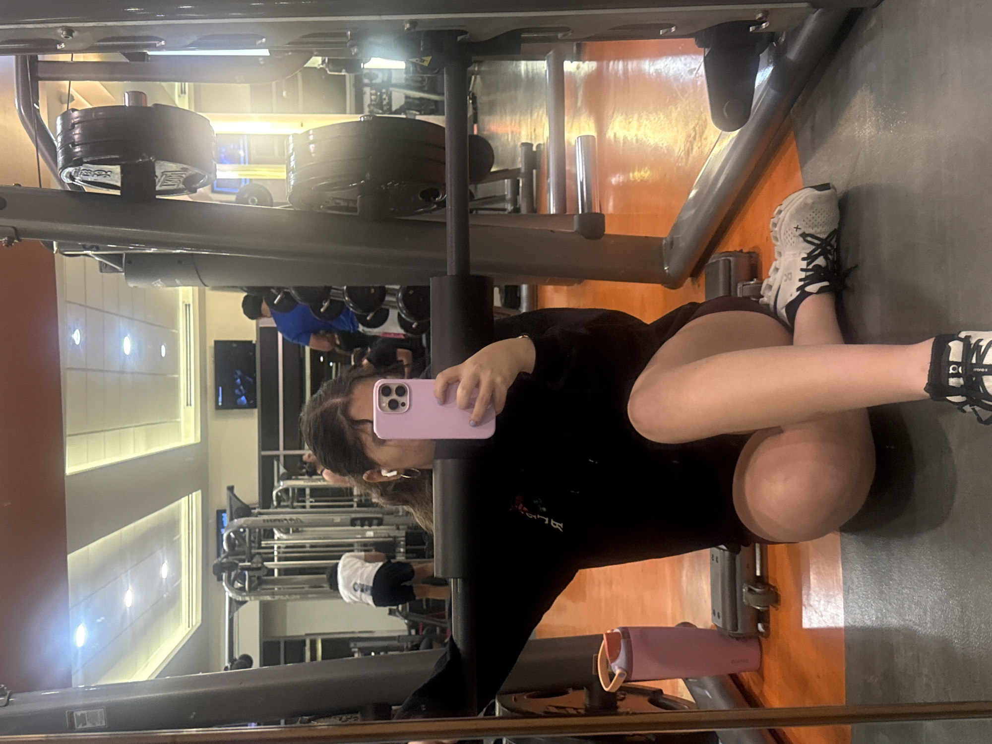 A female subject is depicted seated on the floor of a gymnasium, capturing a selfie using a light purple smartphone. She is reflected in a large mirror, which serves as the primary perspective of the image. The subject wears a black long-sleeved top with white text on the left side, dark shorts, and white and black athletic shoes. A white wireless earbud is visible in her right ear, along with a small hoop earring. Her left arm supports her weight against a black padded bar of a gym machine.
The foreground features a dark gray floor section where the subject is seated, transitioning to an orange-brown polished floor further into the background. A light purple water bottle with an orange lid rests on the floor to the subject's left.
The background, visible through the mirror's reflection, displays various gym equipment, including a large multi-station weight machine with prominent black weight plates, one showing "35" and indistinct branding. Other gym users are present: a male in a white t-shirt and dark shorts facing away from the camera, and another male in a blue t-shirt near a weight rack. Overhead recessed lighting illuminates a light-colored ceiling. Two wall-mounted flat-screen televisions are also visible in the reflection. The specific location within Mexico City cannot be determined from the visual information provided.