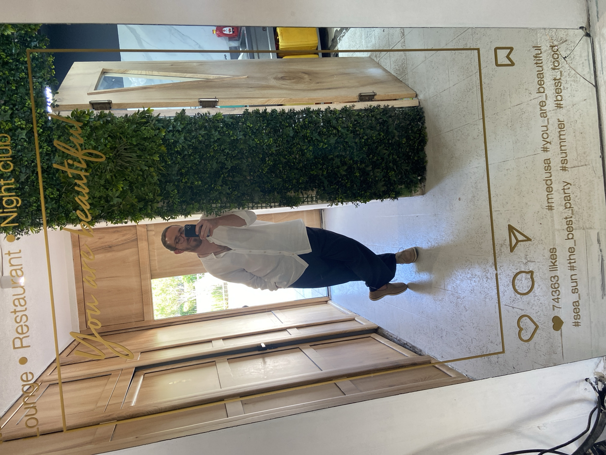 A selfie photograph captures a man standing in front of a mirror adorned with a decorative frame that mimics an Instagram post. The mirror reflects a section of what appears to be the interior of a lounge, restaurant, or night club, indicated by the text on the mirror itself. The man is wearing a white button-down shirt and dark trousers, and he's holding a phone to take the picture, his face visible in the reflection. The mirror's frame includes design elements such as a heart, speech bubble, paper airplane, and square, along with hashtags like "#medusa #you_are_beautiful" and text like "74363 likes." The location is Petrovac centar, Montenegro. The background reflected in the mirror shows elements of interior design, including wooden panels and decorative plants.