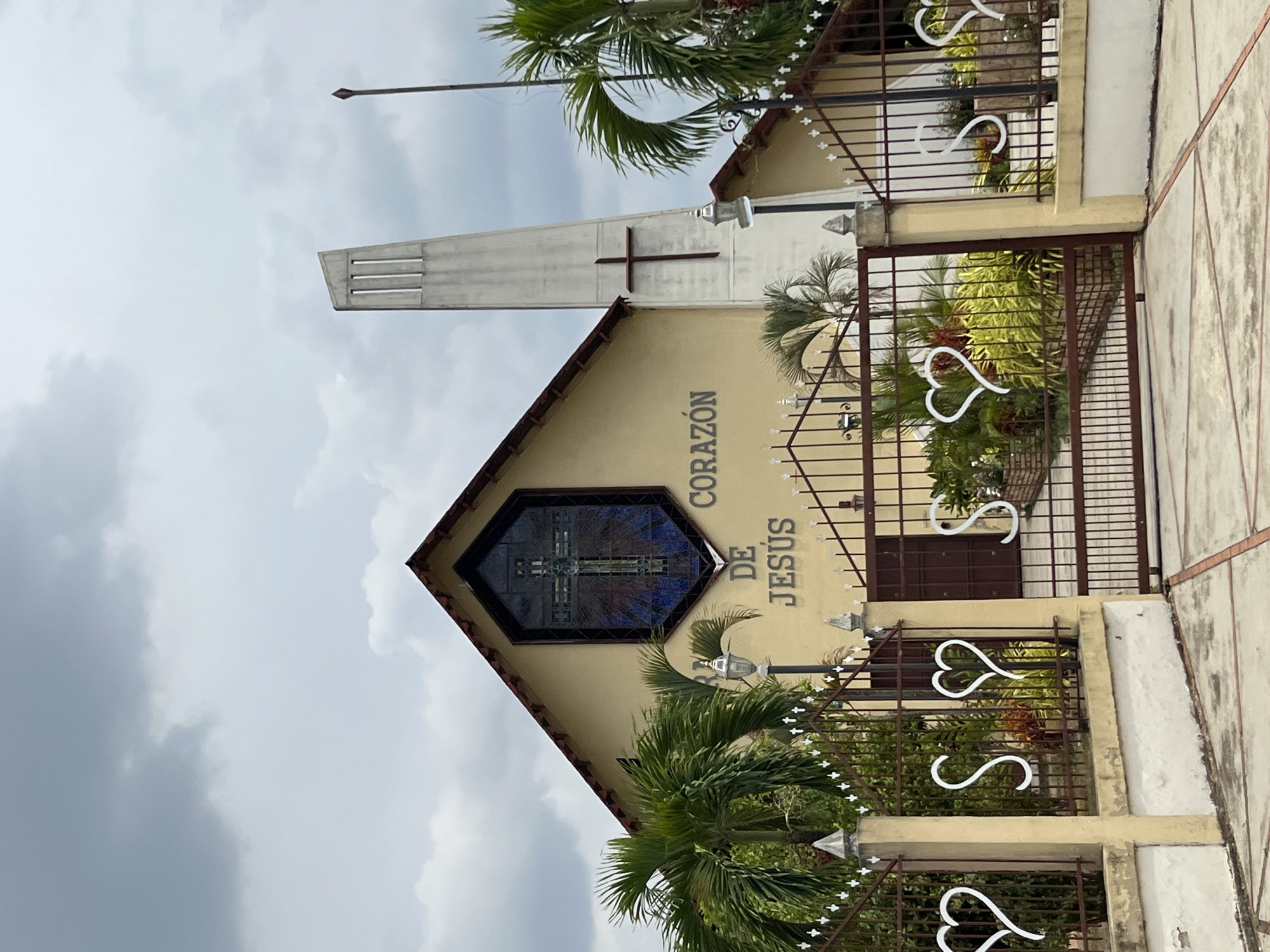 The image shows the "Corazón de Jesús" Catholic Church in Tinaquillo, Venezuela. The church features a tall, concrete tower topped with a cross, attached to a lower, tan-colored building. The building has an octagonal stained-glass window depicting a cross. The church's name is written vertically on the side of the building. A decorative metal fence with heart-shaped accents surrounds the property. The sky is overcast, and there are palm trees in the foreground.