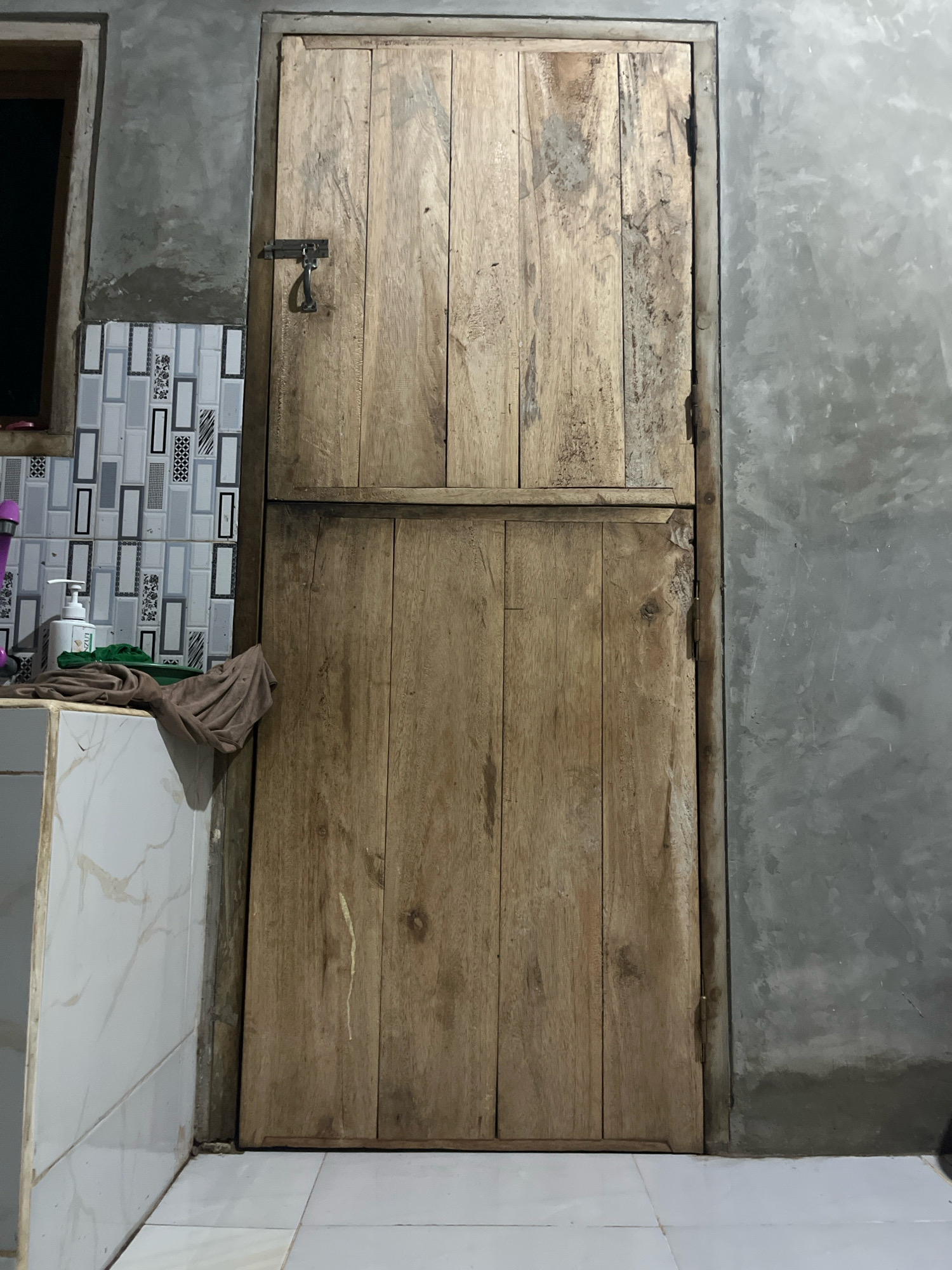 A wooden door, constructed from vertical planks and bisected horizontally by a wooden beam, is shown from a frontal perspective. The door is secured with a metal latch and hinges, and it is set against a textured gray wall. To the left of the door, a tiled wall with a geometric pattern is visible, with a white soap dispenser and folded brown cloth resting on a tiled surface below a small window. The flooring consists of white square tiles. The scene appears to be an interior space, possibly a bathroom or utility area. No people are present in the image. The overall impression is of a utilitarian and somewhat rustic environment.