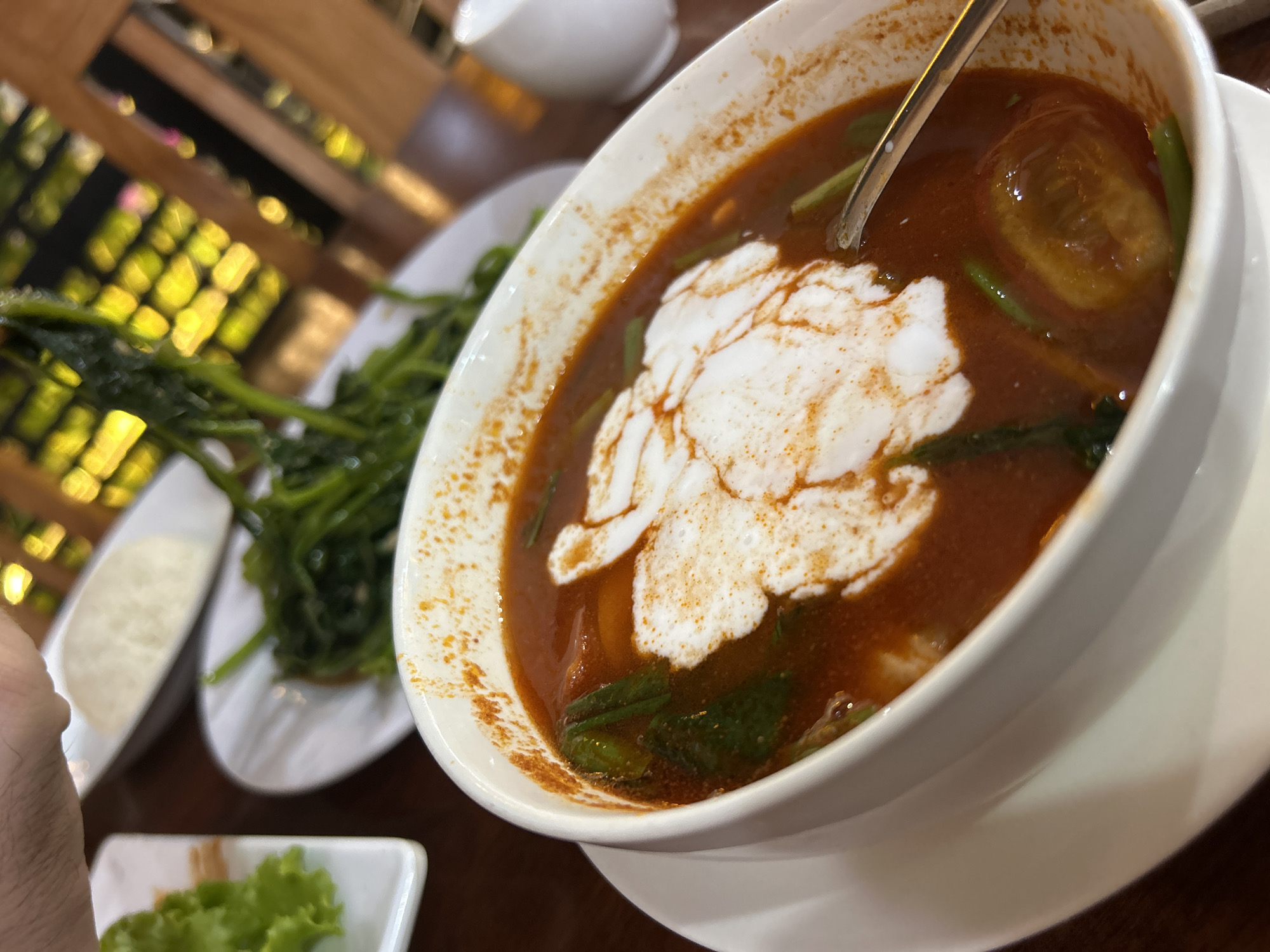 The image shows a bowl of Vietnamese Tom Yum soup, likely in Nha Trang, Vietnam. The soup is served in a white bowl, containing a reddish-brown broth with green vegetables, a slice of tomato, and a swirl of white coconut milk. A spoon is visible inside the bowl. A plate of morning glory is placed on the table beside the soup and other ingredients in white saucers. The setting suggests a restaurant or dining area with a blurred wooden background.