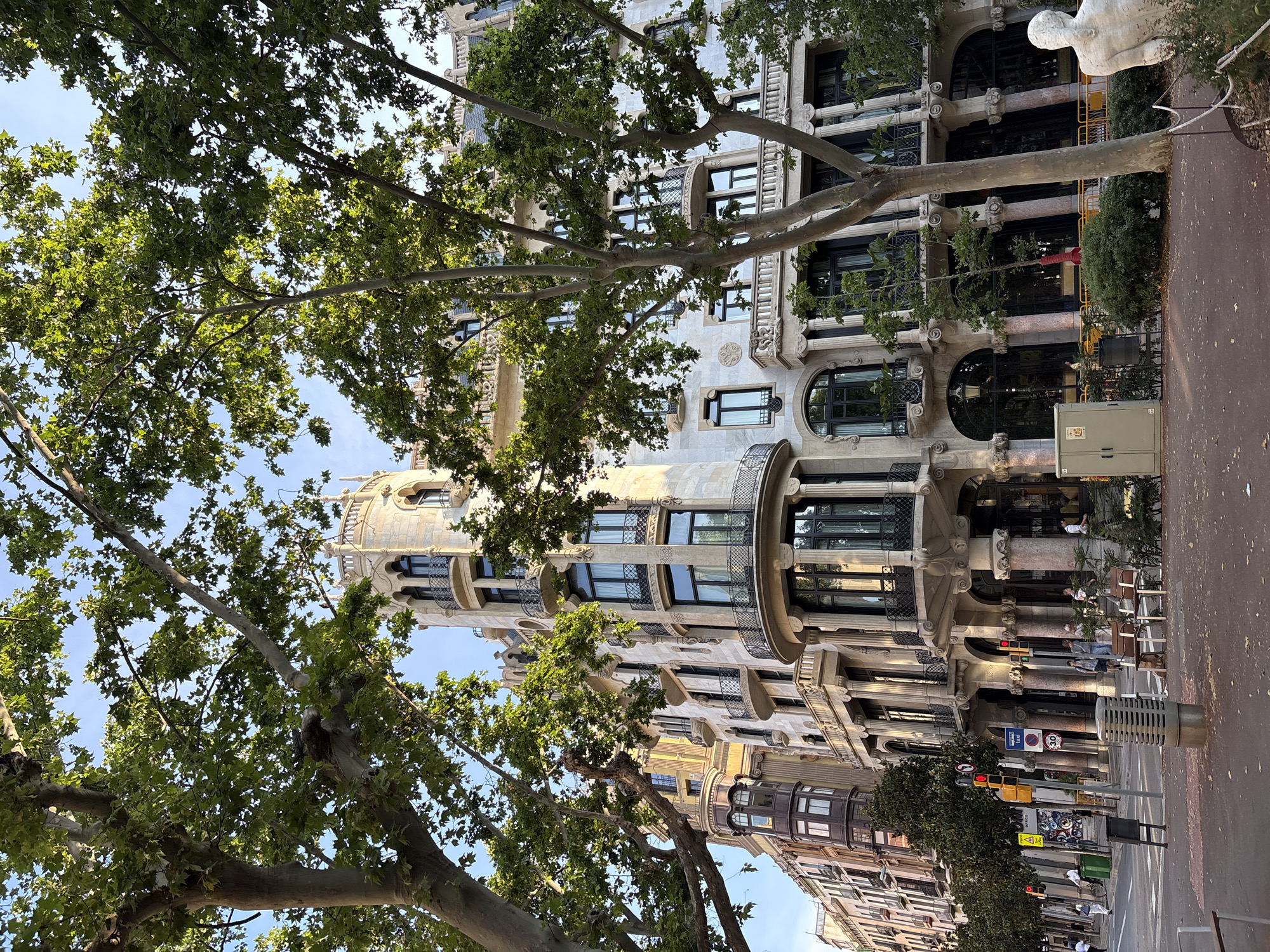 The image shows a partial view of Casa Batlló in Barcelona, Spain, as seen from the street level. The building's distinctive facade, with its unique windows and organic shapes, is partly obscured by trees with green leaves. The street is visible, with pedestrian traffic and signage. A sculpture is visible atop a column on the right-hand side of the image.