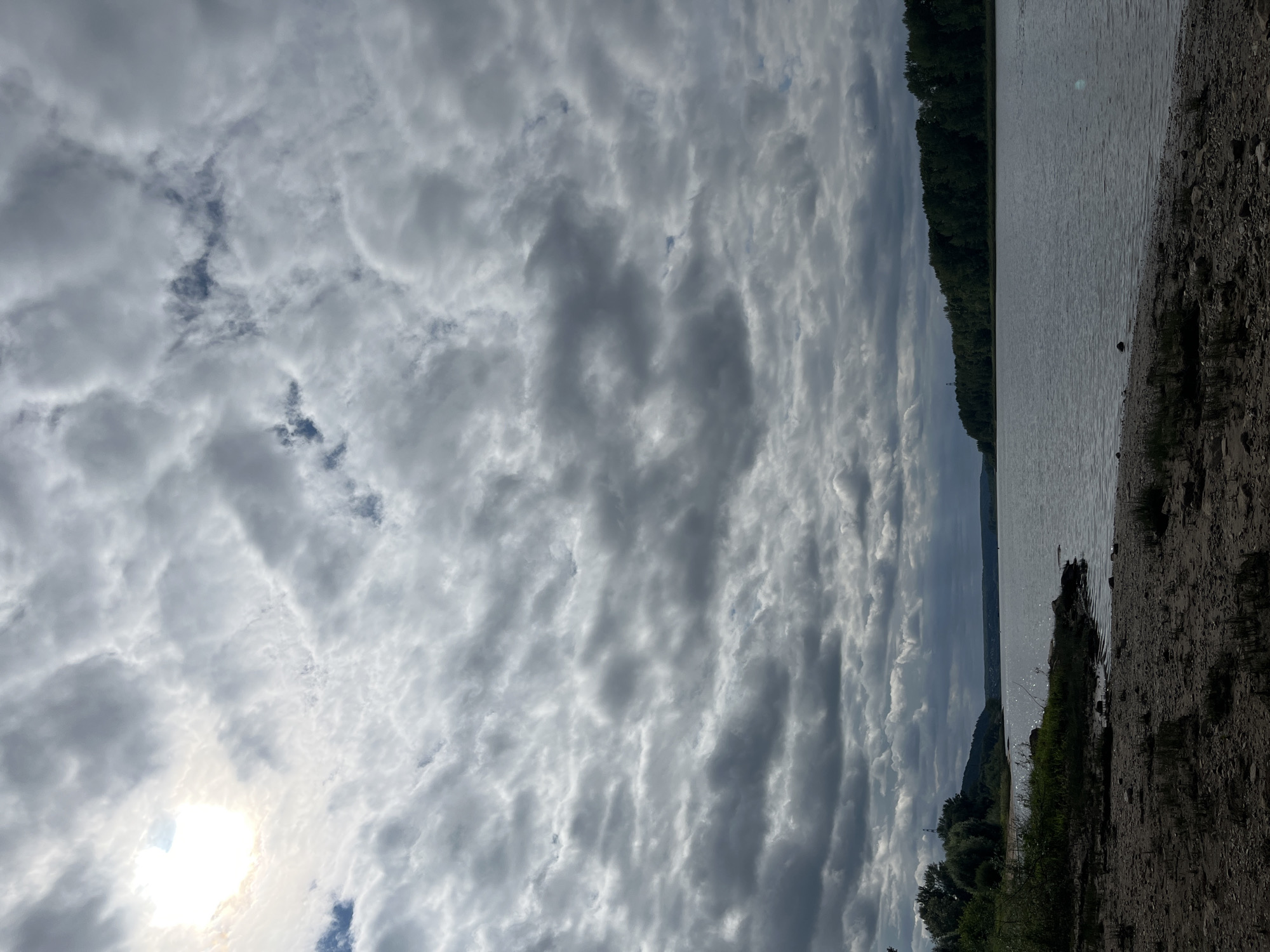 The image depicts a wide, calm river with a sandy shore in the foreground. The shore is sparsely covered with dry grass and small pebbles. The river extends into the distance, bordered by a dense line of dark green trees on the right bank and a hill with trees on the left bank. The sky is heavily overcast with dramatic, layered grey clouds, and a bright sun is visible as a luminous disc peeking through a gap in the clouds in the upper left corner, casting a soft glow. The overall atmosphere is subdued and somewhat dramatic due to the cloudy sky. The location is identified as the Kama River near the city of Chaikovsky, Russia. There are no people or visible actions taking place in the scene.