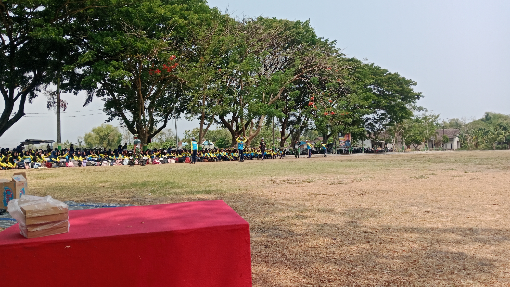 The image depicts an outdoor gathering on a dry grass field, possibly a schoolyard or park, in Madiun, Indonesia. A large group of people, predominantly young individuals wearing yellow shirts and headscarves, are seated on the ground in neat rows under the shade of mature trees. Several individuals in military-style uniforms are present, standing and observing the seated group. In the foreground, a vibrant red platform or stage is visible, with a stack of boxed items covered in plastic wrap placed on it. Beyond the seated crowd, there are buildings, parked motorcycles, and a billboard with images and text, suggesting a public event or activity. The sky is clear and pale blue, indicating a sunny day.