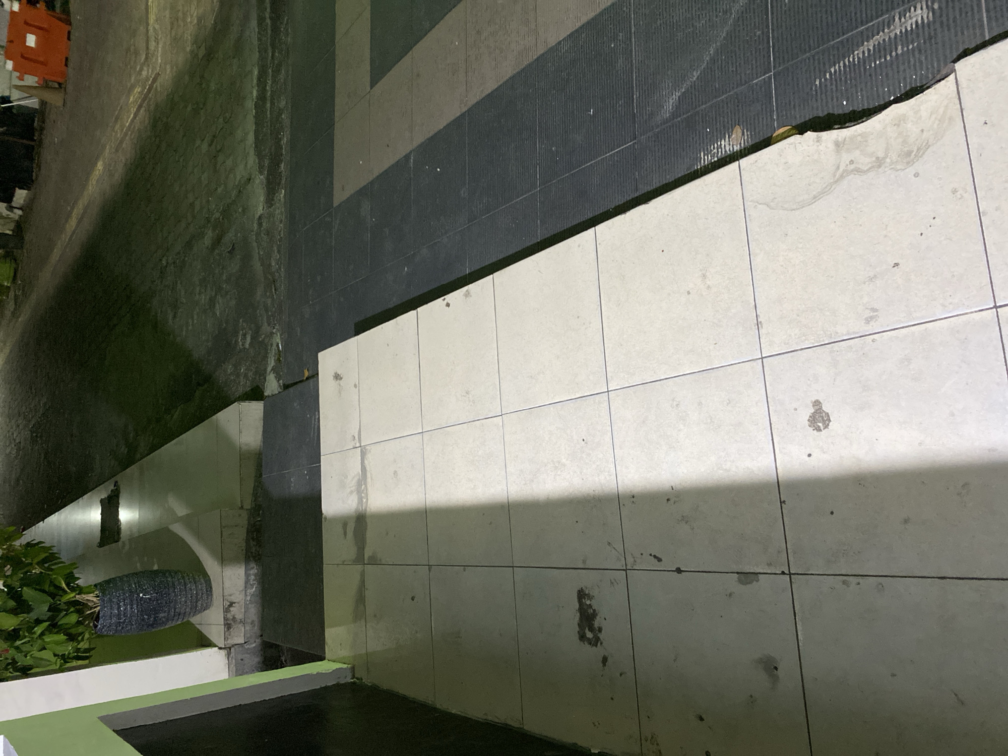 The image shows a tiled walkway at night. The tiles are light-colored and appear somewhat dirty, with some dark stains visible. A shadow is cast diagonally across the tiles from the left side of the frame. To the right of the light tiles, there is a section of darker, textured tiles. In the upper left portion of the image, a dark-colored planter filled with green foliage is visible against a green wall and a white pillar. The background shows a paved area with a light-colored stripe, illuminated by artificial light. Orange construction barriers are visible in the far background, along with other dark shapes that are indistinct. The scene appears to be an outdoor area, possibly a pathway or entrance to a building.