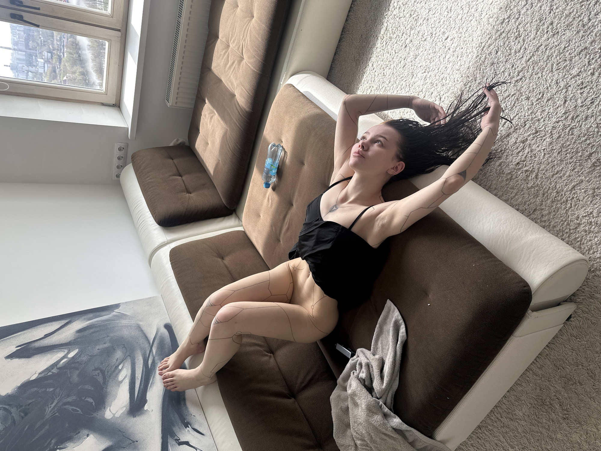 A young woman is depicted reclining diagonally on a brown and white sectional sofa in an indoor setting. She is positioned on her back, looking upwards with a contemplative expression. Her dark hair appears wet and is slightly disheveled, with her left hand grasping strands of it. She wears a black strappy top, and her lower body is partially nude, revealing intricate black geometric lines drawn onto her skin, resembling body art, visible on her arms, torso, and legs.
To her right, on the sofa cushion, there is a clear plastic bottle of water with blue labeling, partially visible as "БонАква" (BonAqua). A light grey towel is draped over the sofa near her feet.
The room is brightly lit by natural light streaming through a large window on the left side of the frame. The window offers a view of an urban or suburban landscape, including buildings and some greenery, consistent with the provided location context of Khimki, Russia. Below the window are white walls and three electrical outlets. The floor is covered with a light-colored, shaggy carpet. In the bottom left corner, a large abstract artwork in shades of grey and white lies on the floor. The scene conveys a sense of relaxation, possibly after a shower, in a sunlit modern interior.