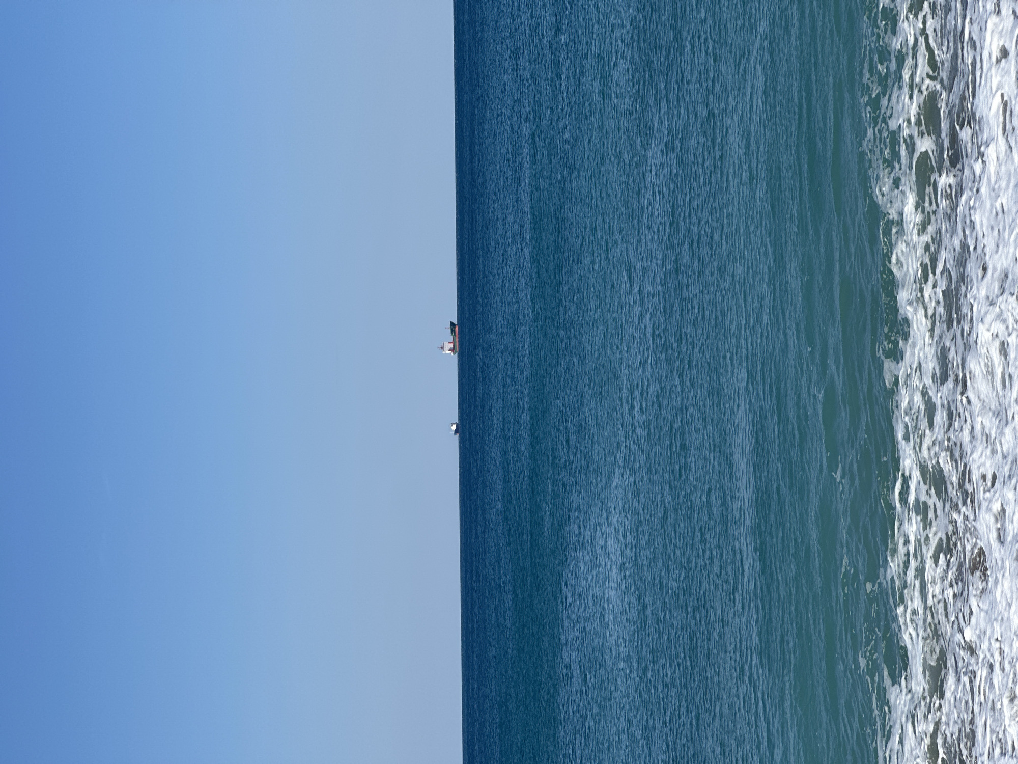 The image depicts a view of the sea near Šušanj, Montenegro. The scene is split between a clear blue sky on the left and the dark blue water on the right. A small boat is visible in the distance near the center of the image. White frothy waves are crashing along the right edge of the image.