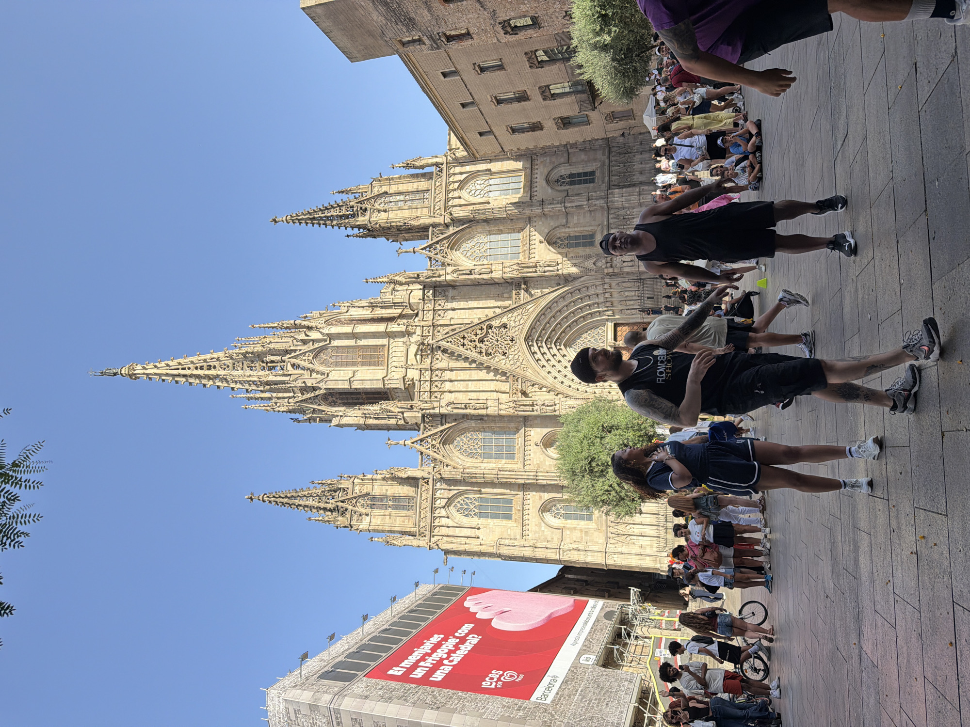This image shows a wide shot of the Barcelona Cathedral in Barcelona, Spain, on a clear, sunny day. The cathedral's impressive Gothic architecture dominates the background, with its spires, intricate carvings, and large arched windows clearly visible. In the foreground, a crowd of people are gathered in the plaza. Some are standing and talking, while others are sitting on the ground or walking around. A large red billboard is displayed on the left side of the image, advertising for "Locos por Barcelona." The ground is paved with stone tiles. The overall atmosphere appears to be lively and bustling, with tourists and locals enjoying the public space.
