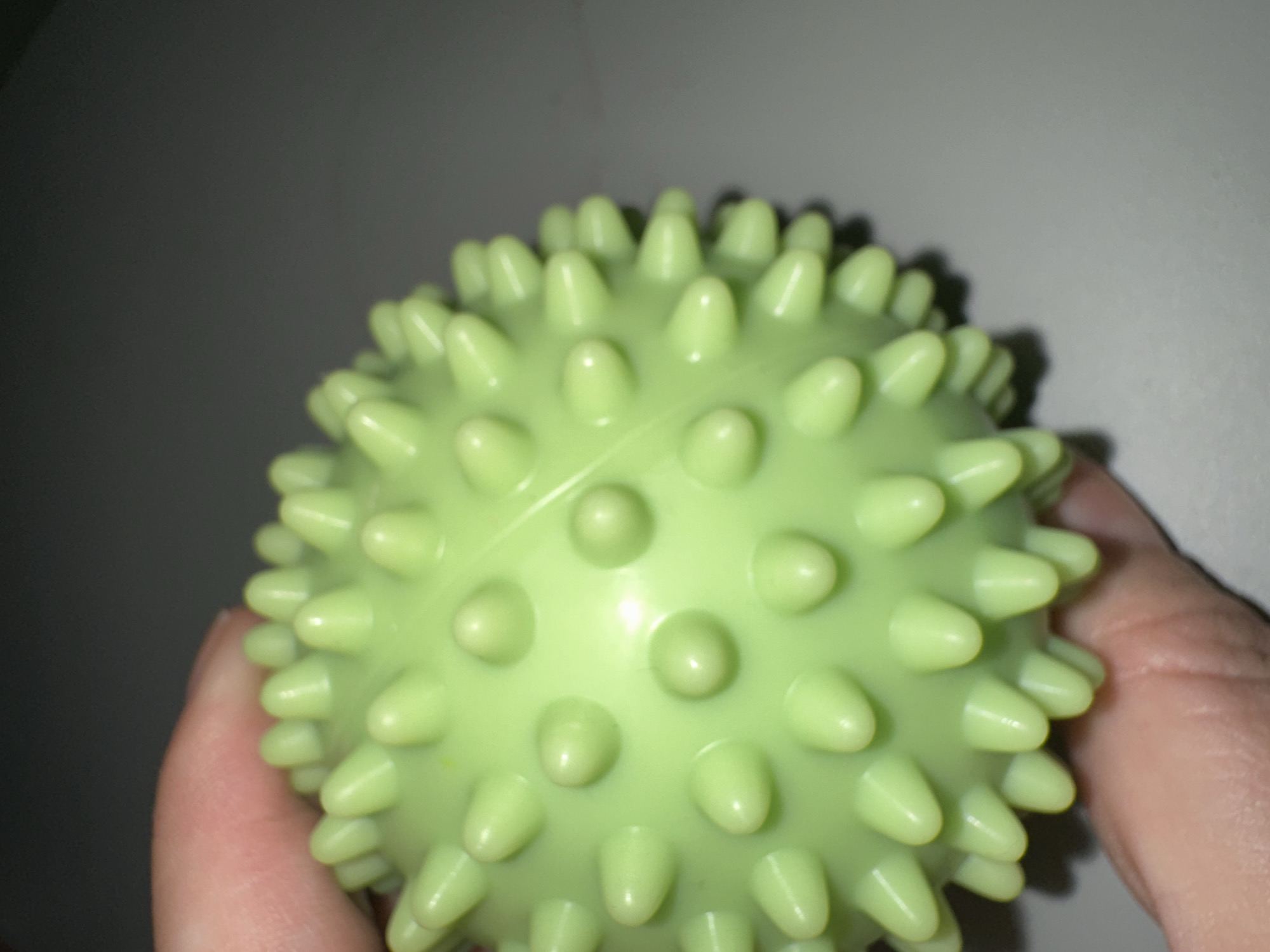 A light green, spiky sensory ball is being held by a human hand. The ball is roughly spherical and covered in numerous conical protrusions. The background is dark and out of focus, with a subtle gray surface visible in the lower right quadrant. The image appears to be a close-up shot, likely taken indoors. There are no identifiable landmarks or context to place this image in Krefeld, Germany.