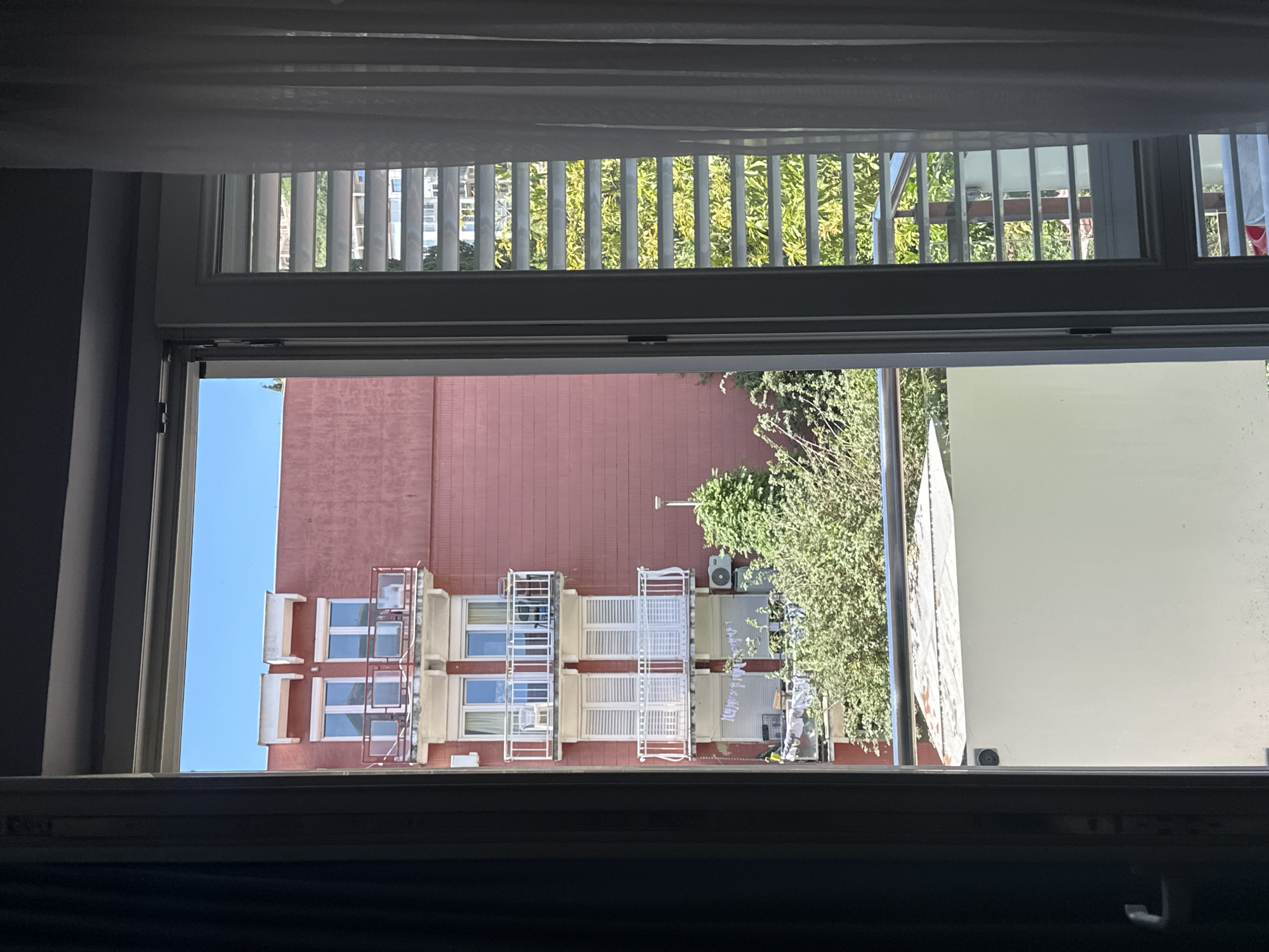 View from an open window in Budva, Montenegro. The window looks out onto a multi-story brick building with white balconies and windows. A tree is partially obscuring the view to the right. Above the open window is another window with white blinds. The sky is visible in the open window. The room the window is in is dark.