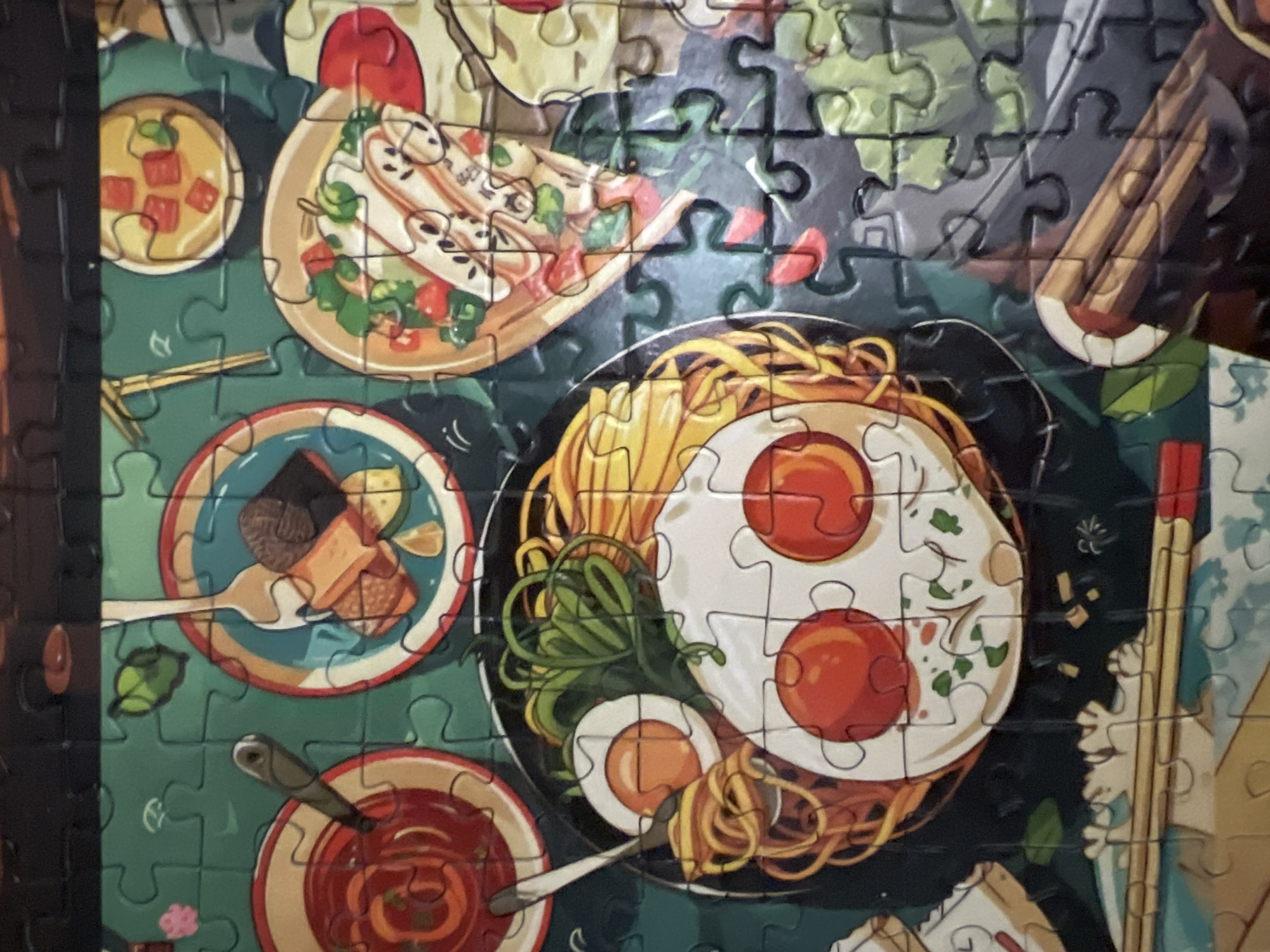 The image is a close-up, overhead shot of a jigsaw puzzle depicting an assortment of Japanese food dishes arranged on a dark green, textured surface. The puzzle pieces are clearly visible, creating a fragmented view of the artwork.
The dishes include:
* A large bowl of ramen with noodles, broth, green onions, and two fried eggs with visible yolks.
* A smaller bowl with a red, sauce-like substance, possibly a dipping sauce, with a spoon in it.
* Another small bowl containing what appears to be cubed pieces of food in a yellow broth.
* A shallow dish with sliced food, possibly sushi or sashimi, topped with green garnishes and red circular pieces.
* A bowl with sliced ingredients and a white base, which could be a type of salad or another appetizer.
* A cylindrical object made of stacked wood-like pieces, possibly chopsticks or a serving utensil.
The overall aesthetic is colorful and illustrative, with bold outlines and bright hues.
There is no indication of the city of Volgograd, Russia, or any other specific geographical location in the image. The scene is purely a depiction of food items on a puzzle.