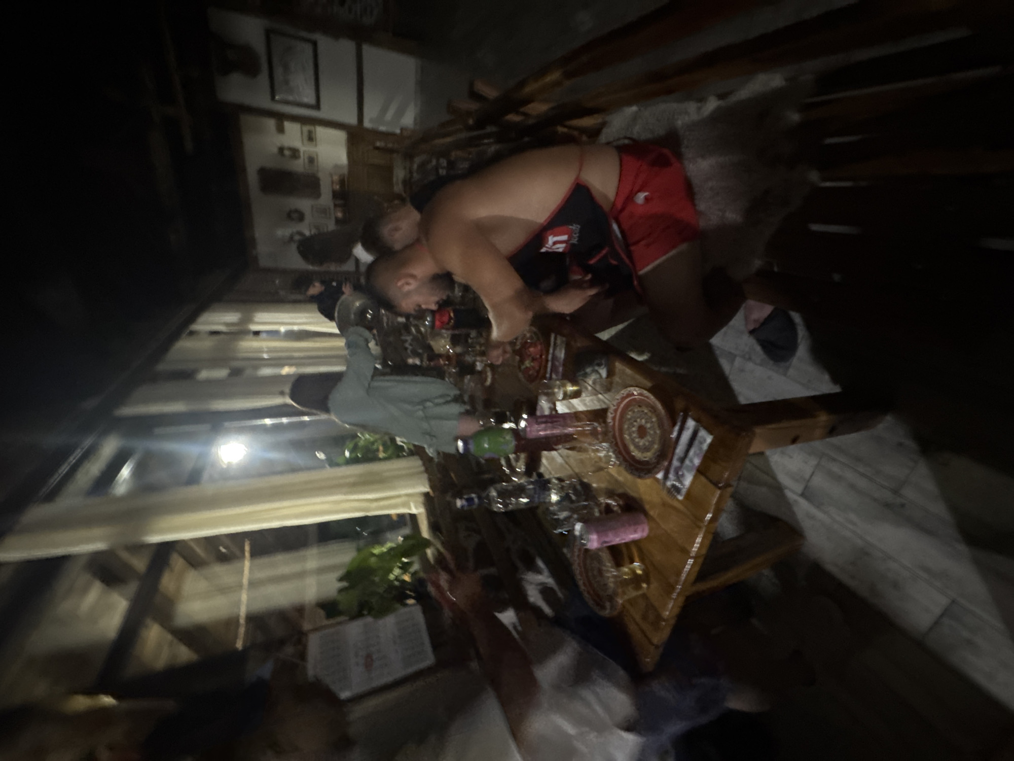 The image depicts a dimly lit, indoor gathering, likely at a restaurant or private residence in Konare, Bulgaria. A group of individuals is seated around a wooden table laden with plates, glasses, and bottles, suggesting a meal or social event.
In the foreground, a man is prominently featured, shirtless and wearing red athletic shorts and a black apron with red trim. He is seated at the table, appearing to be eating or interacting with food. To his left, another person is partially visible, also seated at the table.
Further back, a woman in a light-colored garment is standing and appears to be interacting with someone behind the table. Other individuals are blurred and less distinct, suggesting movement or being out of focus.
The environment features wooden architectural elements, possibly exposed beams or paneling, and large windows with sheer curtains. The lighting is atmospheric, with artificial light sources casting shadows and illuminating parts of the scene. Decorative elements, such as framed pictures on the wall in the background, are also visible. The overall impression is of a casual and convivial gathering.