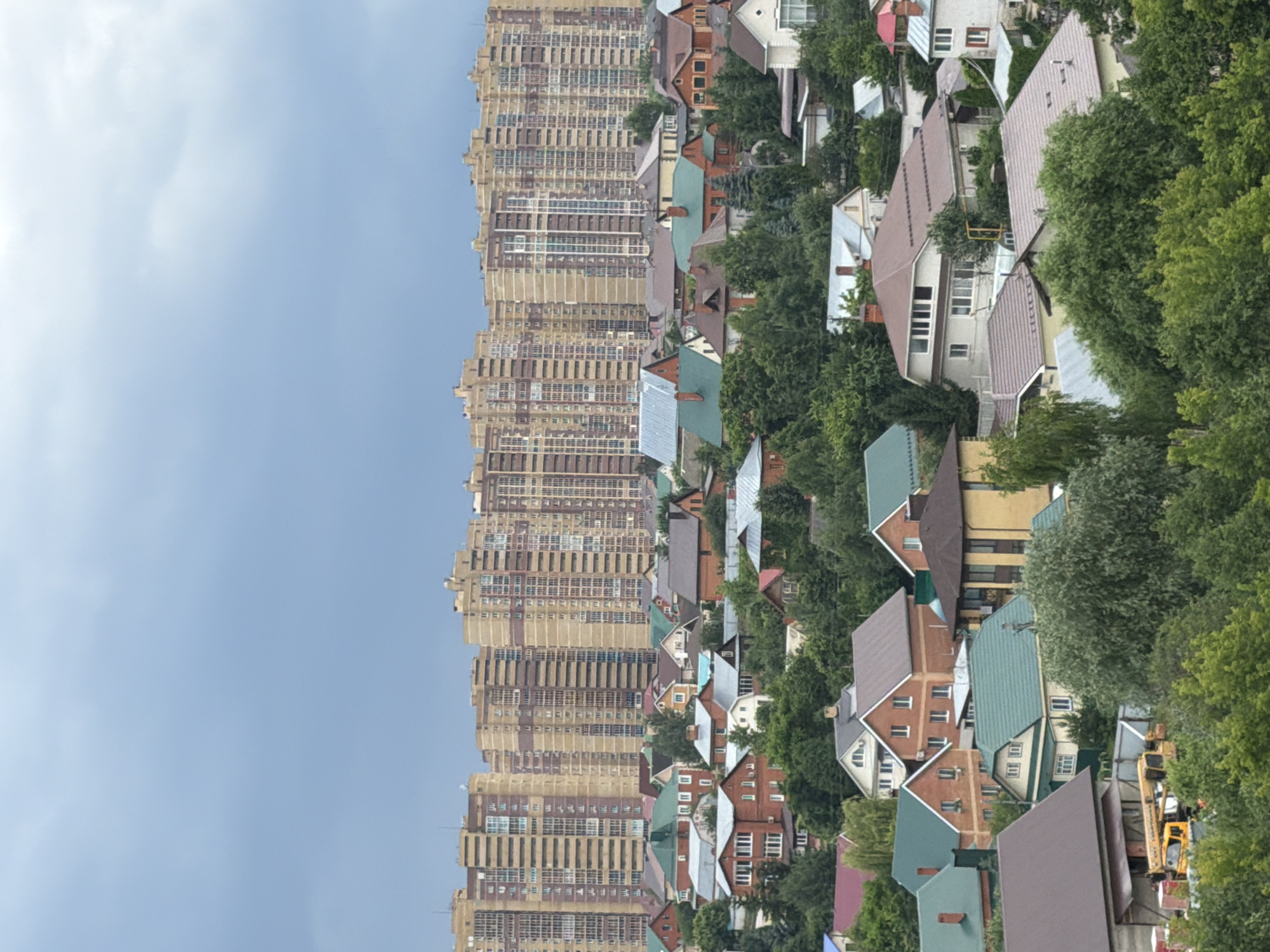 The image shows an aerial view of a suburban area with numerous houses and a backdrop of tall apartment buildings. The houses are of various sizes and architectural styles, mostly featuring pitched roofs. Lush green trees are interspersed throughout the residential area, suggesting a densely vegetated landscape. In the foreground, a yellow construction vehicle, possibly a crane or lift, is visible near a building under construction, with stacks of bricks nearby. The sky is overcast with dark clouds, indicating a possibly stormy or cloudy day. The apartment buildings in the background are uniformly designed, featuring multiple floors and numerous windows, suggesting a modern, densely populated urban development contrasting with the lower-rise residential area in the foreground.