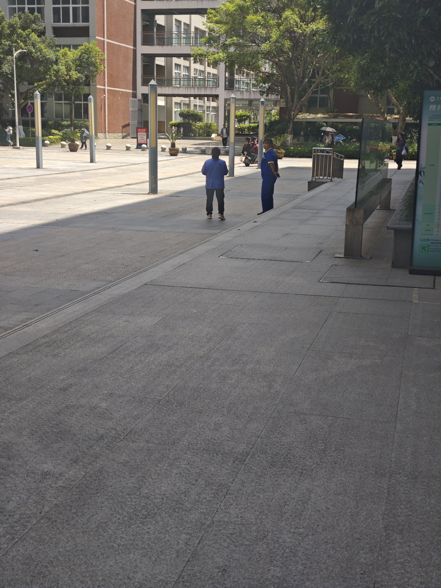 The image depicts an outdoor scene in Chongqing High-tech Zone, China. Two individuals are visible: one in a blue shirt facing away from the camera, and another in a blue uniform. The scene also shows a paved plaza with trees, poles, and buildings in the background. There's a bus stop with signage near the right edge of the frame. Other people are present in the background, some walking and one on a scooter. The environment is daytime with strong sunlight casting shadows across the plaza.