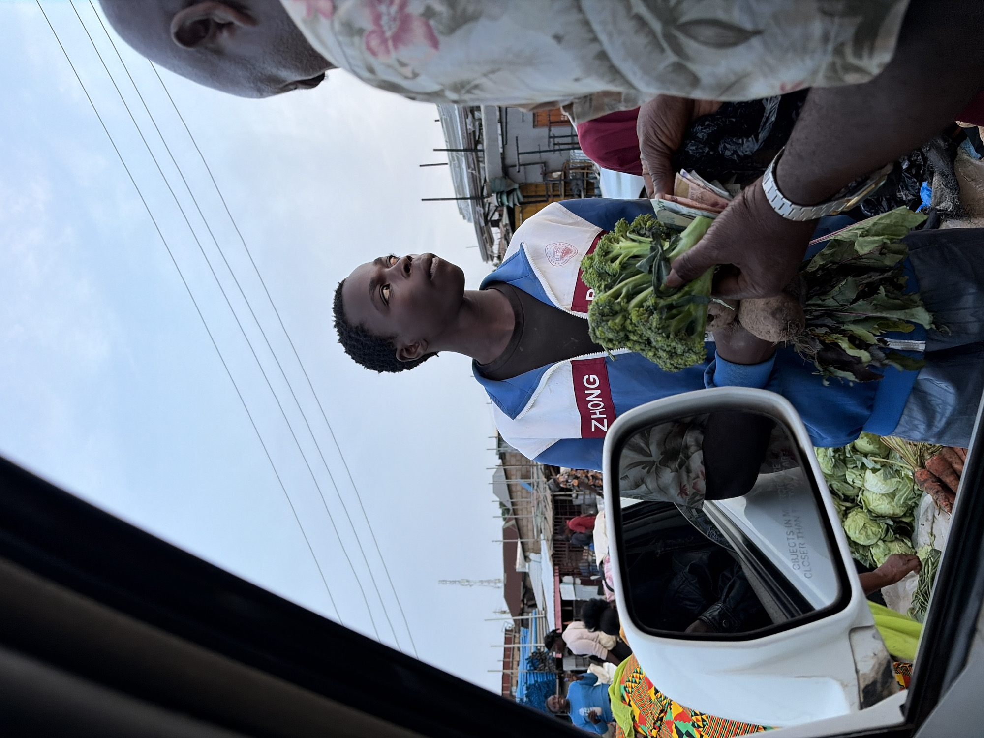 The scene depicts a street market in Gura Top, Nigeria. A young Black male wearing a blue and white jacket with the word "ZHONG" visible stands in the middle of the frame, looking upwards. He appears to be a vendor or a customer. An older person with a flowered shirt is visible in the top left corner. An arm wearing a metal watch is holding cash in one hand and a bunch of broccoli and beetroot in the other hand. Cabbages and carrots are also visible in the bottom right. In the background, market stalls, a telecom tower, and other people are visible. The image appears to have been taken from inside a car, with the car window frame and side mirror visible on the left side of the frame, reflecting the inside of the car.