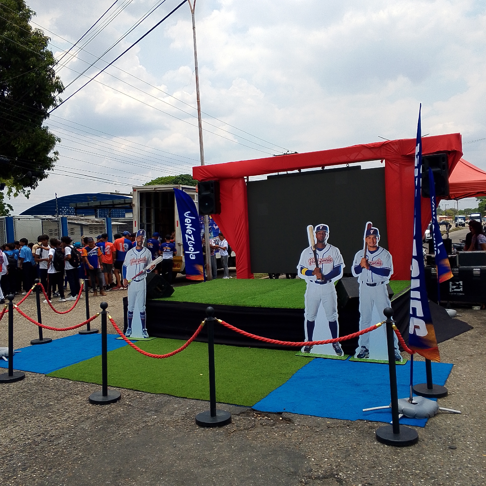 The image shows an outdoor promotional event in San Carlos, Venezuela. A small stage is set up with a red frame around a large screen. Two life-size cardboard cutouts of baseball players in Venezuelan national team uniforms stand in front of the stage. A blue and orange flag with "Venezuela" printed on it is positioned on the left side of the stage. A red rope barrier is set up on green and blue artificial turf to create a VIP area in front of the stage. A group of people, mostly young adults, are gathered behind the rope barrier. In the background, a white truck is parked near the stage. A large, blue and white building is visible behind the truck. Power lines are visible overhead against a cloudy sky.