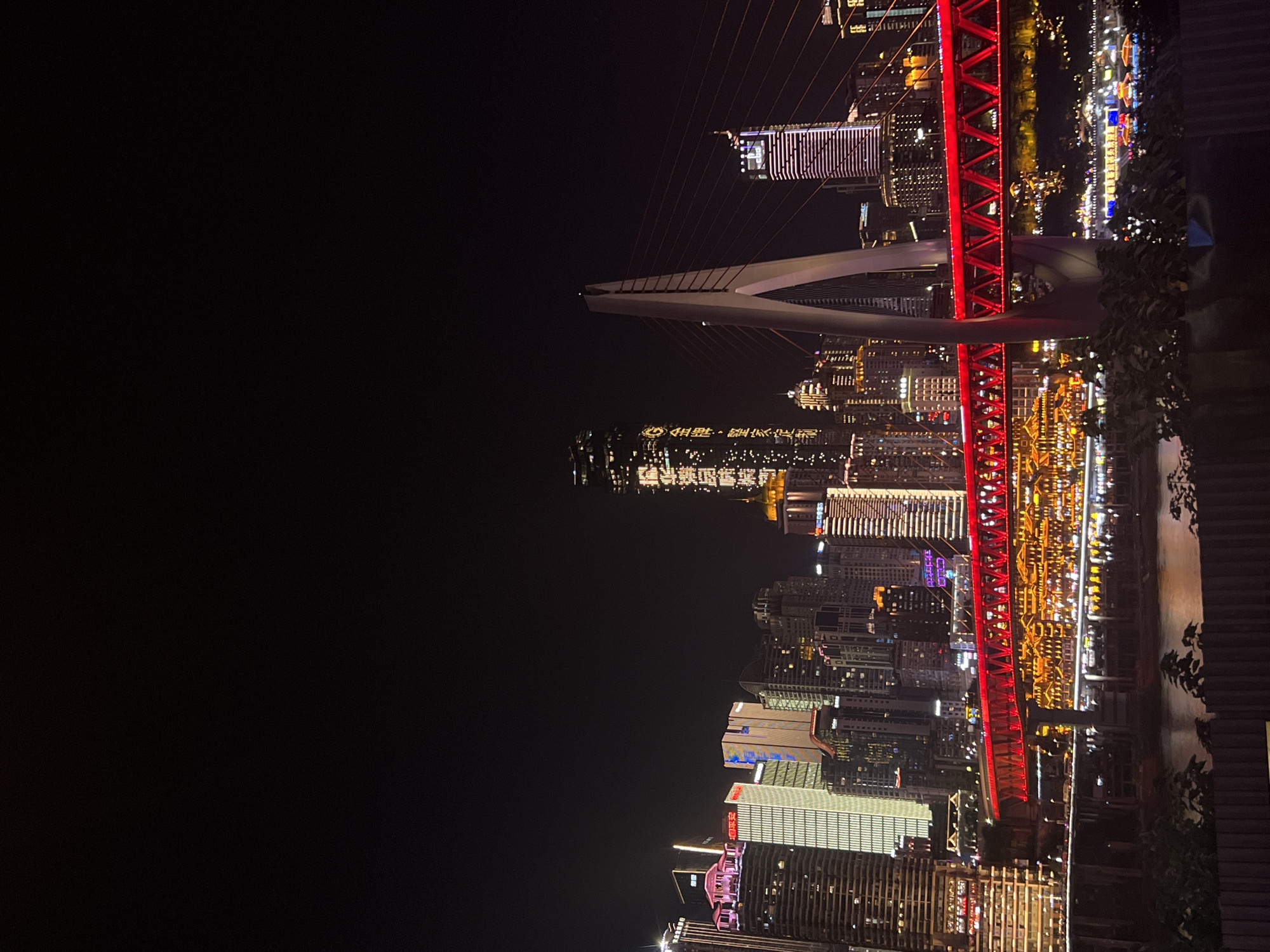 The image captures a nighttime urban landscape in Jiangbeicheng Subdistrict, China, dominated by illuminated skyscrapers and a prominent red bridge. The Hongya Dong area is visible along the waterfront, with its distinctive traditional architecture illuminated by numerous yellow lights. A modern cable-stayed bridge with a white pylon and red support cables spans the river, its red deck lights creating a vibrant contrast against the dark sky. Several high-rise buildings with various architectural styles and lighting schemes are clustered together. One building on the right features vertical pink and white lights, while another on the far left has pink lights atop its structure. The overall scene depicts a bustling cityscape at night, with reflections of the lights visible on the water's surface. The foreground includes a dark fence and some foliage, suggesting a vantage point from a slightly elevated position.