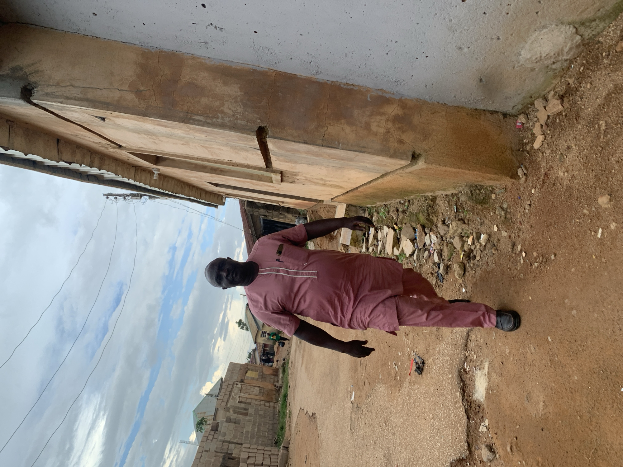 A man walks on a dusty, unpaved road in Jos, Nigeria, wearing a rose-colored traditional outfit. He is the central subject of the image, positioned in the foreground and facing the camera with a neutral expression. The road is in disrepair, with visible potholes and debris scattered along its edges. To the right of the man, a light brown, textured building with a corrugated metal roof stands tall, appearing aged and somewhat weathered. The building has a series of rectangular windows, some of which are open. To the left of the man, a partially constructed brick building and other structures are visible in the background, suggesting a developing urban area. The sky above is overcast with dramatic clouds. The overall scene depicts a common streetscape in Jos, characterized by its unpaved roads, traditional architecture, and bustling, yet humble, urban environment.