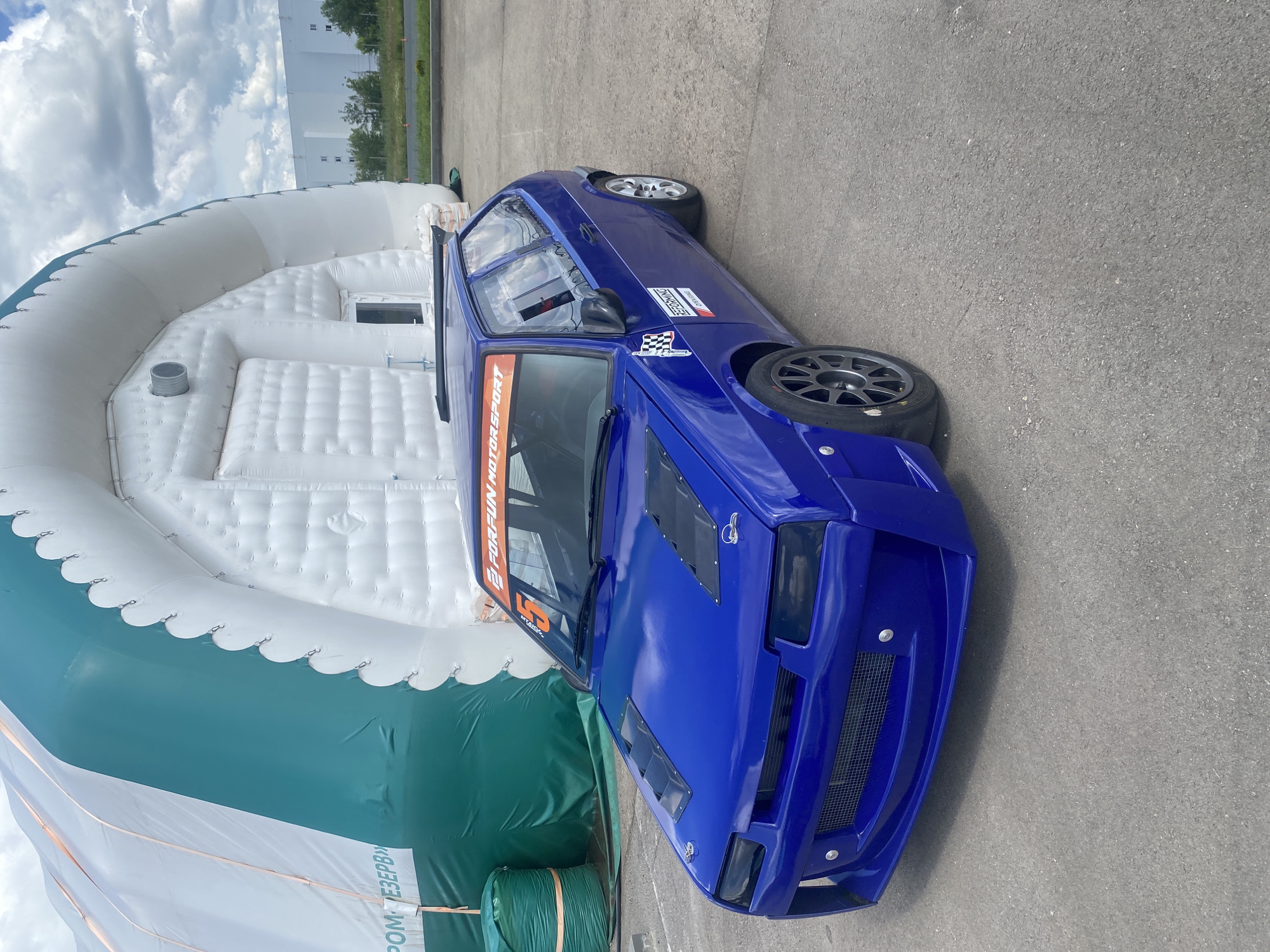 A blue, modified hatchback race car is parked on an asphalt surface in front of a large, white inflatable structure. The car has a custom body kit, including a prominent front spoiler with a mesh grille and several vents on the hood. A small spoiler is visible on the roof. Racing decals are present on the car, including a checkered flag emblem and text that reads "FORFUN MOTORSPORT" on the windshield banner and "REFORMING" on the side. A red number "5" is also visible on the windshield. In the background, a large inflatable structure, predominantly white with a teal green section on the left, dominates the scene. Partially visible to the right of the inflatable structure is a long, white industrial building. The sky above is partly cloudy. The location is identified as Svetloe Pole, Russia.