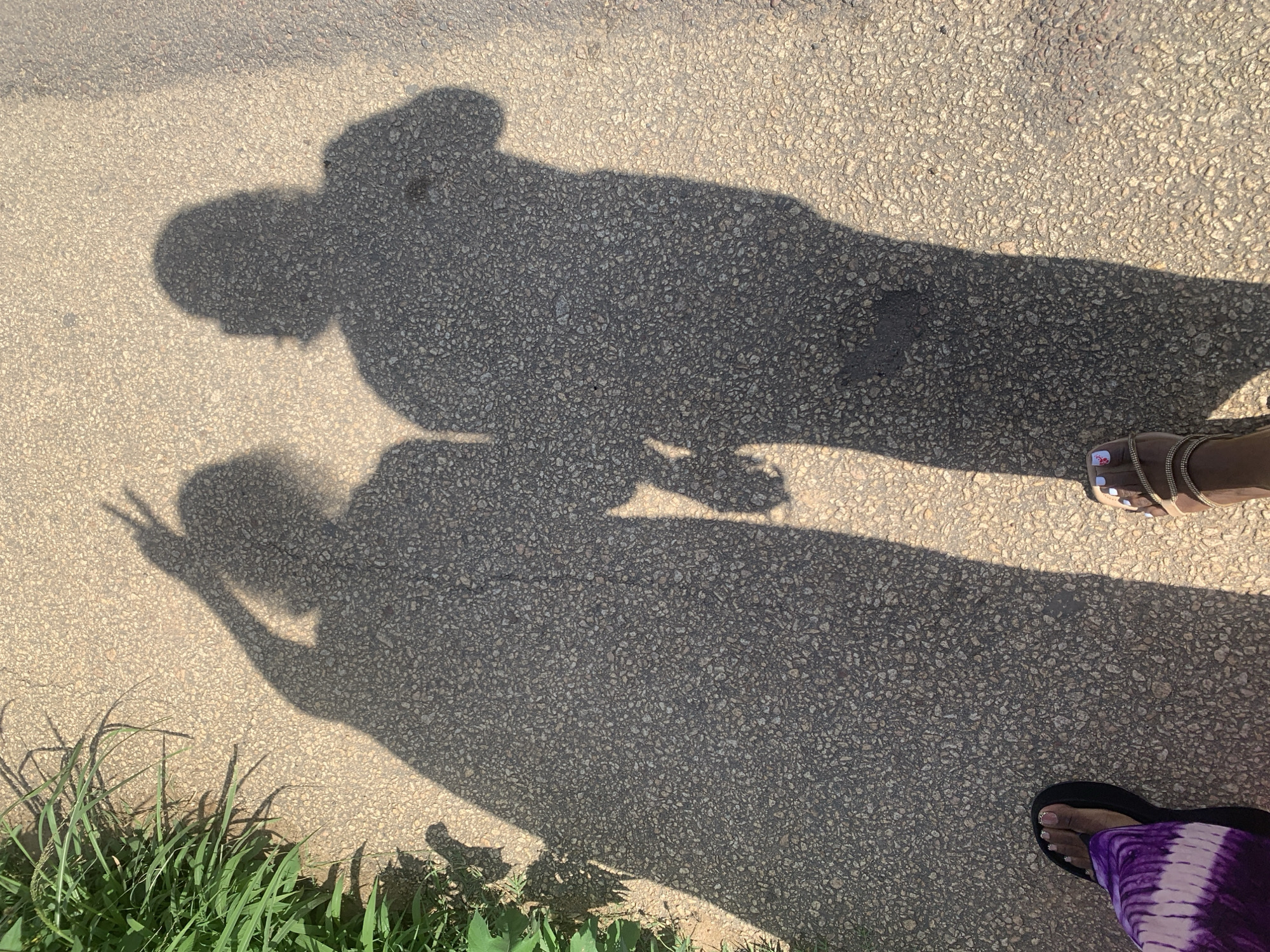 Two individuals' shadows are cast on a paved surface, likely a sidewalk or path, with a section of green vegetation on the left side of the frame. The shadow on the left displays a hand raised with two fingers extended in a peace sign. The shadow on the right appears to be of a person standing close to the first, with their hands clasped or arms linked. The feet of the two individuals are visible in the foreground; one is wearing a purple and white tie-dye garment and a black sandal, while the other is wearing a tan sandal with decorative straps. The paved surface is textured with small stones. The scene suggests an outdoor setting, possibly a casual stroll. This image could have been taken in Rukuba Road, Nigeria, given the environmental context and typical urban features.