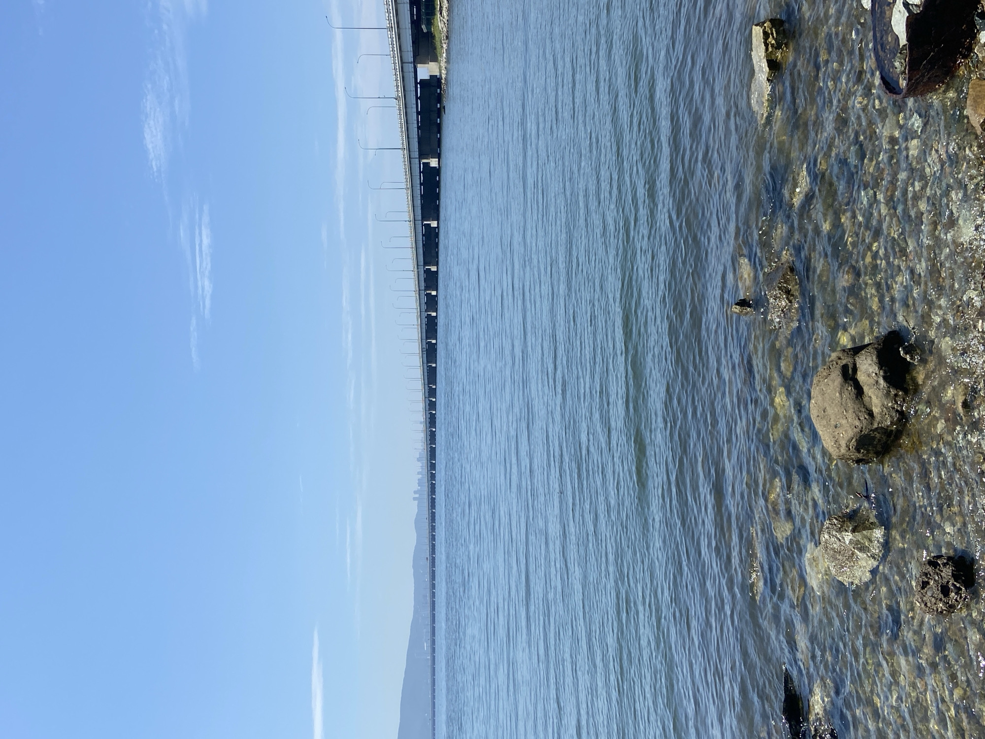 The image depicts a wide, calm body of water, likely a bay or strait, under a clear blue sky with scattered thin clouds. A long, multi-span bridge stretches across the water in the mid-ground, connecting distant landmasses. The bridge is supported by numerous concrete pillars and features lampposts lining its edges. On the far shore, a city skyline with tall buildings is visible, nestled against rolling hills. The foreground shows a rocky shoreline with clear, shallow water revealing submerged pebbles and a few larger rocks, including a dark, roughly spherical rock. To the right in the very foreground, a rusted metal object, possibly a barrel or container, is partially visible. The scene appears tranquil and captures a view of a coastal urban landscape. Given the description of a long bridge connecting landmasses and a city skyline with hills visible in the distance, this is likely the Zolotoy Rog Bay Bridge (Golden Horn Bay Bridge) in Vladivostok, Russia, which connects the city center to the Churkin Peninsula.