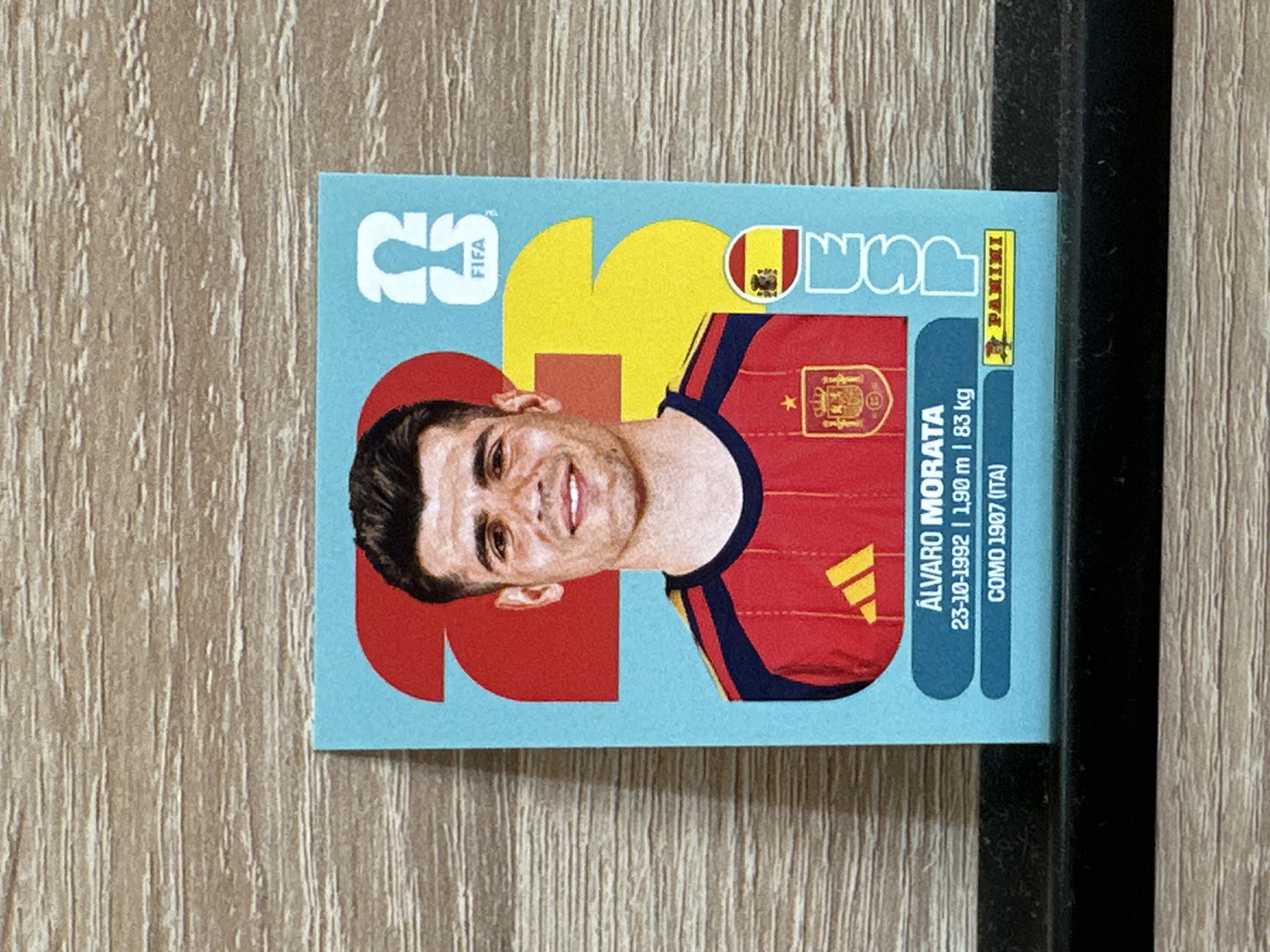 The image shows a close-up, slightly angled view of a Panini FIFA World Cup sticker featuring Álvaro Morata, a Spanish footballer. The sticker is light blue with large red and yellow graphic elements. Morata is depicted from the chest up, wearing a red Spanish national team jersey with a dark blue trim on the collar and shoulders. His jersey has the Spanish crest and an Adidas logo. The sticker includes text below his image: "ÁLVARO MORATA 23-10-1992 | 1.90 m | 83 kg COMO 1907 (ITA)" and a Panini logo. The background of the sticker is a light blue color. The sticker is placed on a surface with a visible wooden grain texture, which appears to be a table or shelf. The sticker is slightly tilted. There is no indication of the city of Prisovo, Bulgaria, in the image.