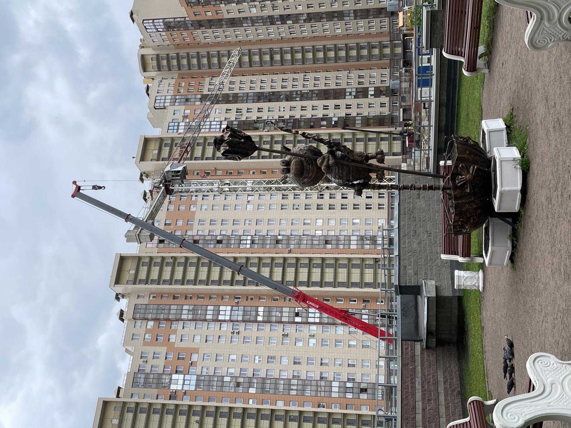 The image shows a modern sculpture in a park setting, set against the backdrop of high-rise apartment buildings, possibly located in a newer residential area of Saint Petersburg, Russia. A large crane, of the type used in building construction, is also present. The foreground contains benches and planters, and a group of pigeons is visible on the ground. The weather appears overcast.