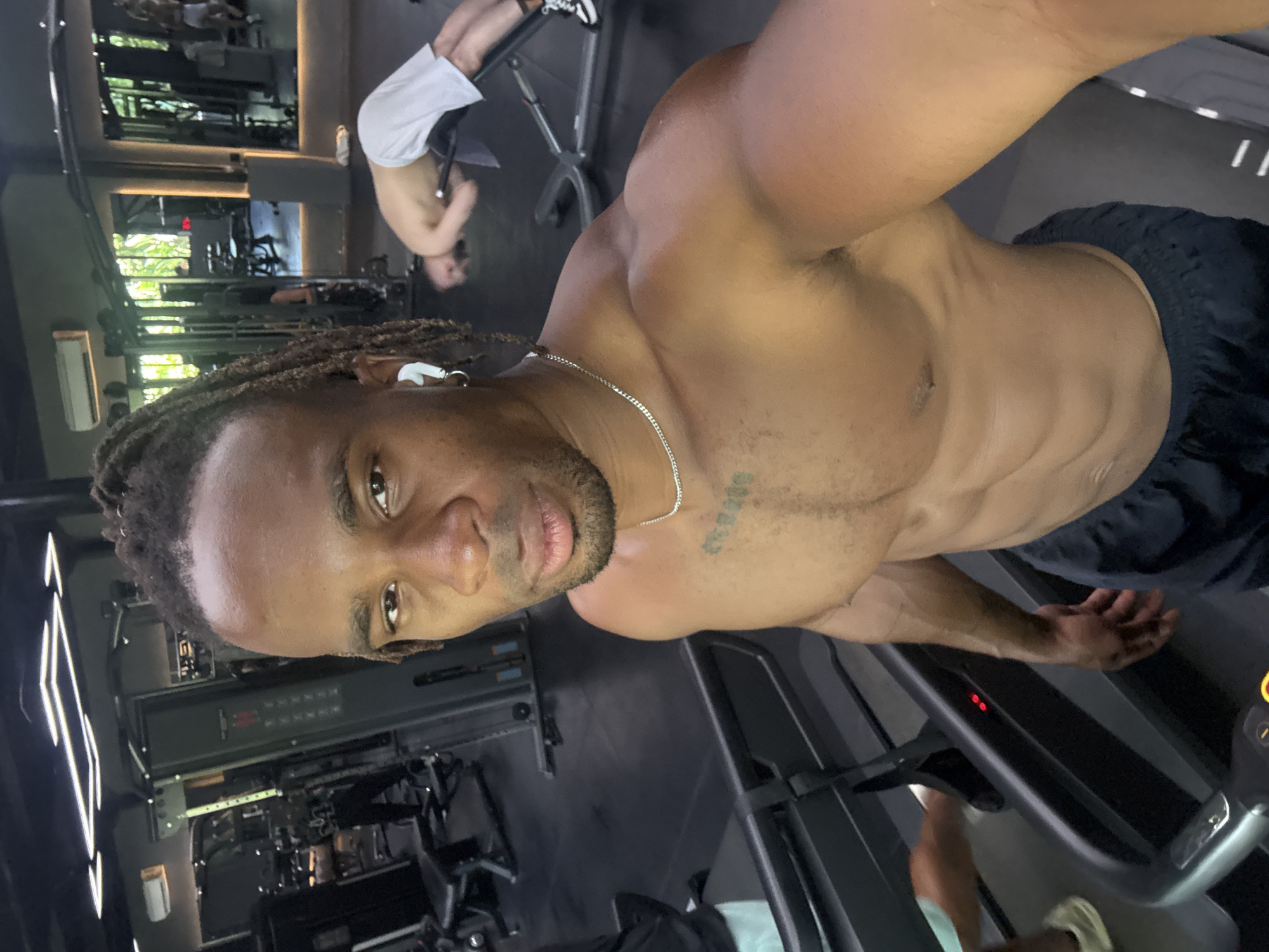 A shirtless man with dreadlocks stands on a treadmill in a gym. He is wearing a silver necklace and has an earbud in his left ear. A tattoo is visible on his chest. Other gym equipment and people are in the background. The gym appears to be located in an unknown location, Unknown.