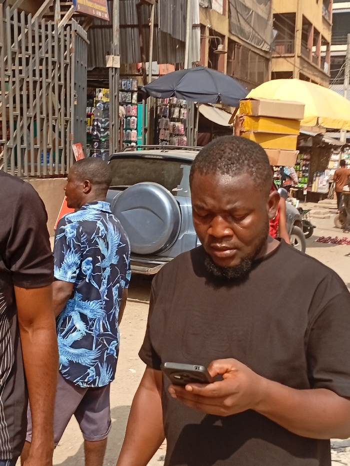 Amidst a bustling urban landscape, a man focused on his smartphone dominates the foreground. He wears a plain black t-shirt. Behind him, a person in a vibrant blue shirt with a bird pattern stands partially visible, and the edge of another person in a striped shirt is also present. A parked car with a spare tire cover, street vendors with umbrellas, and stacked merchandise fill the background, illustrating the dynamic and crowded setting. The scene is captured in natural daylight.