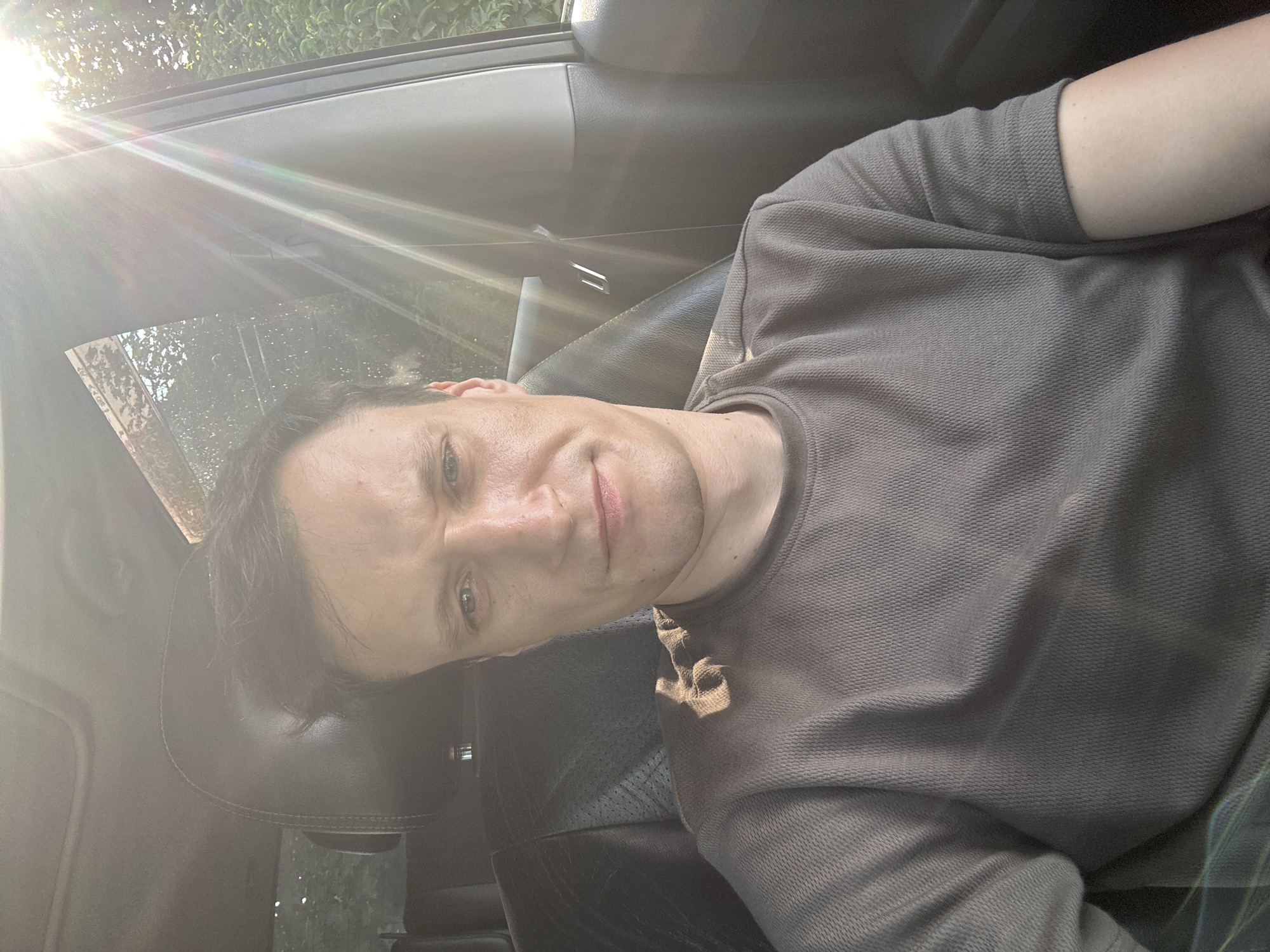 A man is sitting in the driver's seat of a car, with the sun shining through the windshield and side window, creating lens flare. He is wearing a grey t-shirt. The interior of the car is dark leather. The scene appears to be in an outdoor setting, possibly a street or parking area, with greenery visible outside the windows. There is no discernible information about the location within the city of Shatava, Ukraine.