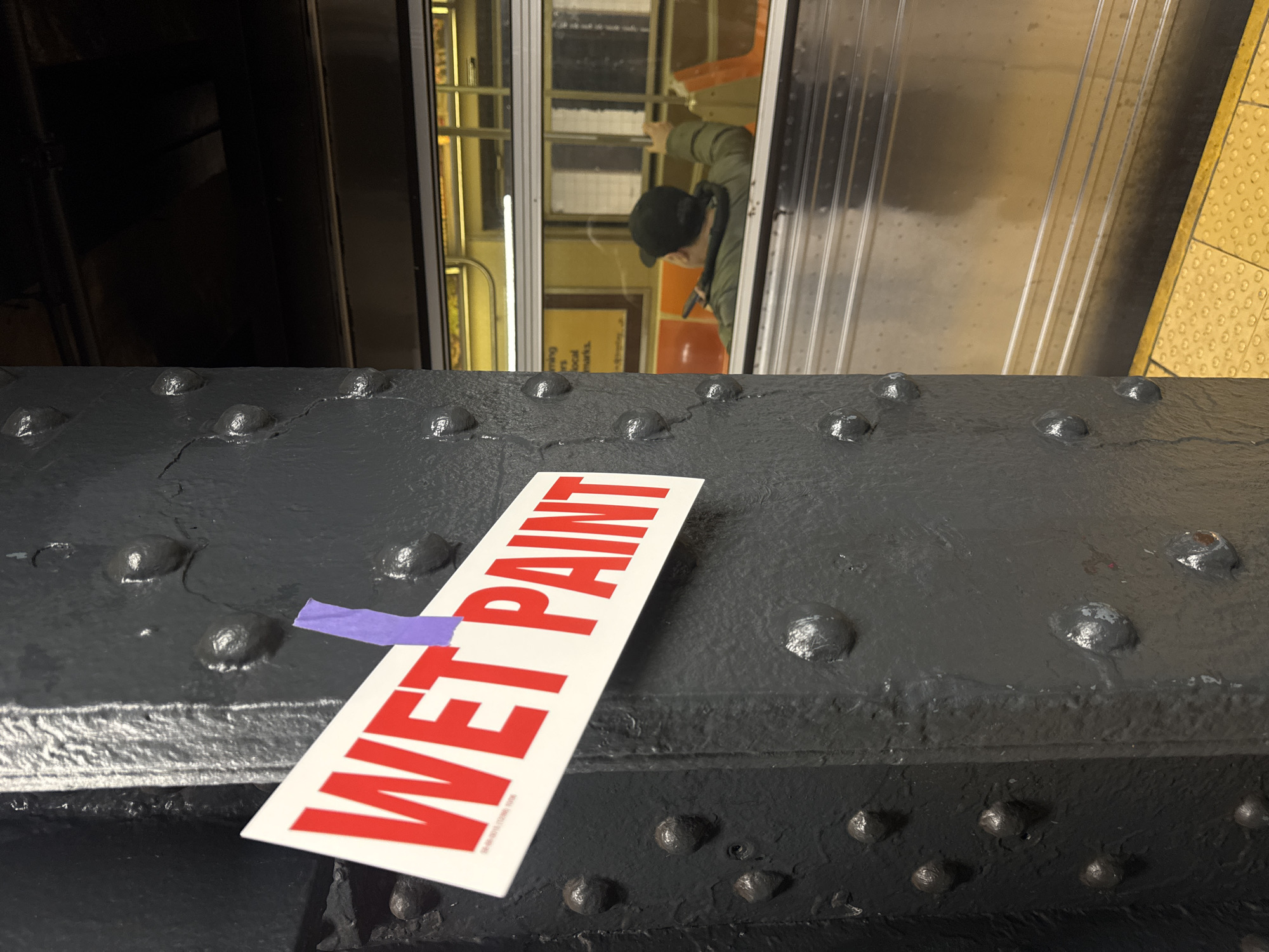 The image depicts a New York City subway station platform and a subway train. In the foreground, a dark grey, heavily riveted metal structural column is positioned on the left. A white sign with "WET PAINT" in prominent red capital letters is attached to the column with a piece of purple tape; smaller text "SB-S8-0013 (12/06) 100K" is visible at the bottom left of the sign. To the right, a silver New York City subway train is stopped at the platform. Through the train's window, a person is visible from the back, wearing a dark green jacket and a black baseball cap, seated on an orange subway seat. Their right arm is slightly raised, and a dark rectangular object is visible in their left hand, resting on their lap. The train platform features a yellow textured tactile warning strip along the edge. The environment is dimly lit, indicative of an underground setting.