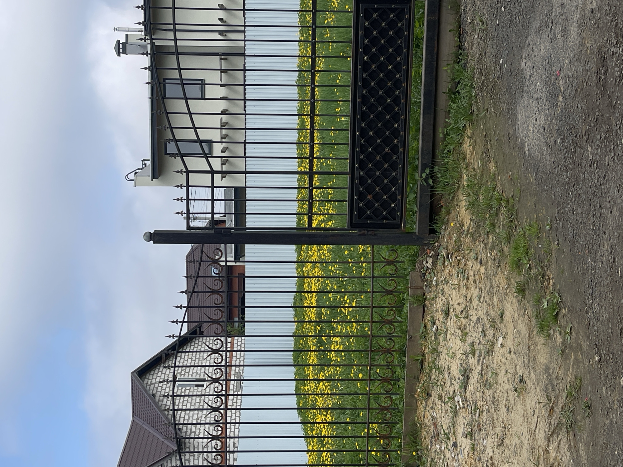 The image shows a wrought-iron fence and gate in Misaylovo, Russia. Through the fence, a field of yellow flowers is visible. Two houses are partially visible through the fence. One house is light-colored with a dark roof and the other is white brick with a brown roof. A dirt and gravel path is visible to the right of the fence. The sky is overcast.