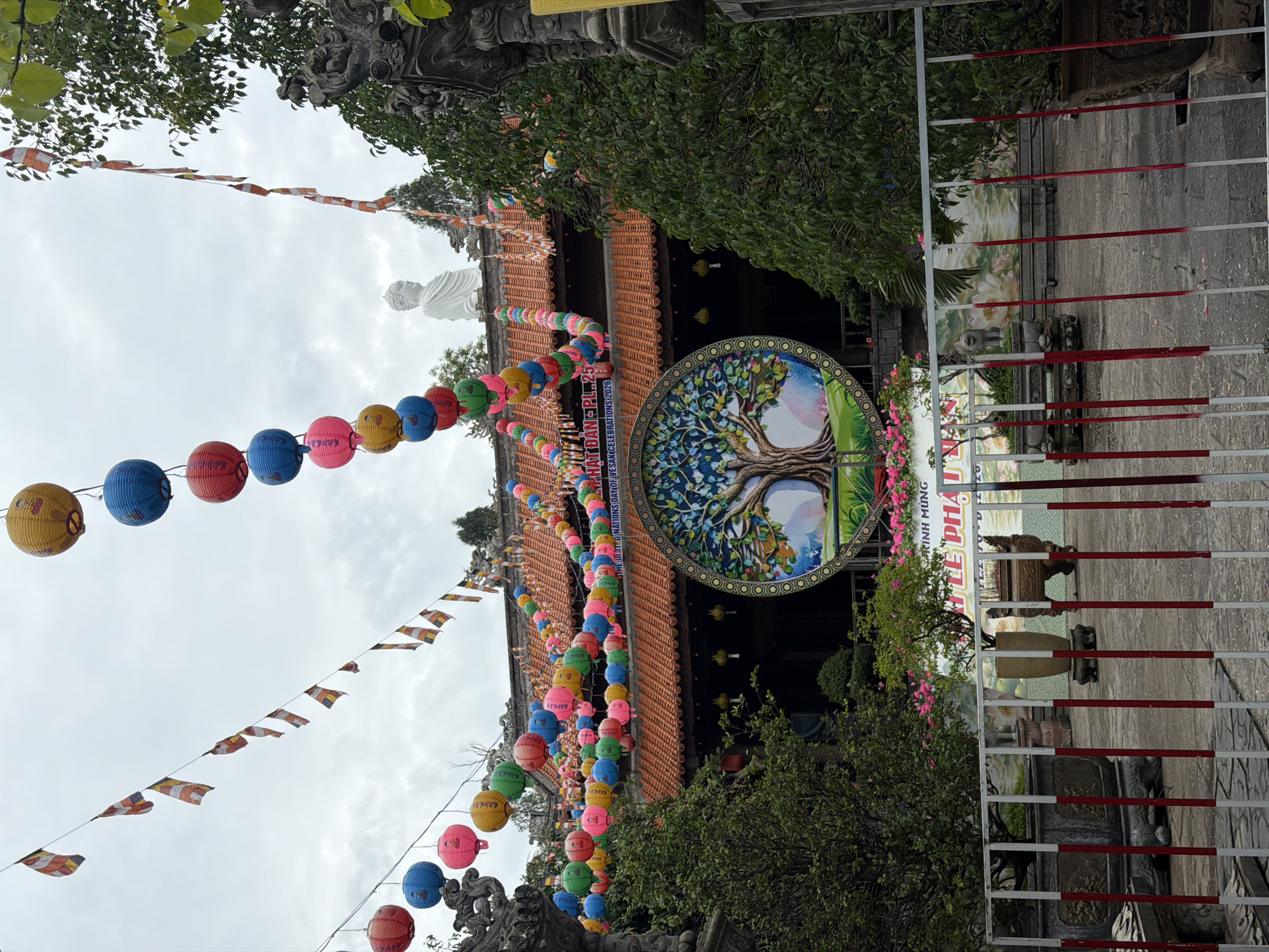 The image depicts a temple in Tay Nha Trang Ward, Vietnam, adorned with colorful lanterns and flags strung across the front of the structure. A round sign with a tree illustration hangs on the front of the temple. A white Buddha statue is visible in the background, atop the temple. A red and white metal fence is in the foreground. The scene appears to be outdoors, possibly during a festival or celebration. The sky is overcast. There is some text present on the temple’s facade: “THE UTM.DINATIONS DAY OF”, “KÍNH MÙNG”, “LE PHA U”, “HAT DAN PL 25”, “NS 2026”.
