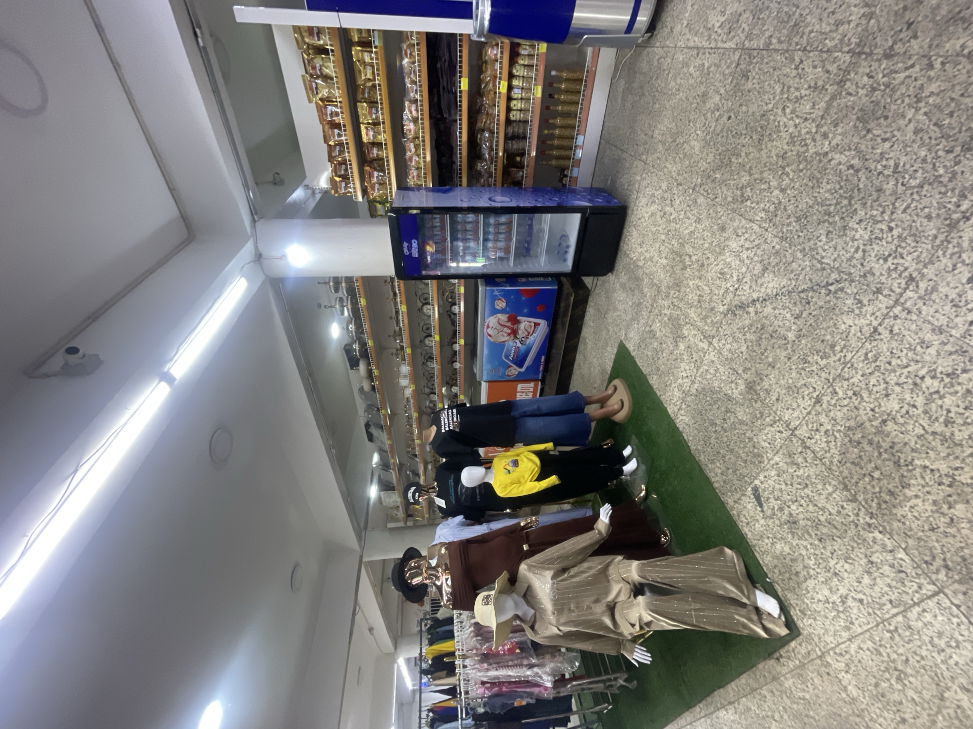 This is a full-shot, eye-level, indoor photograph of a retail store in Galadimawa Village, Nigeria. The scene is lit by fluorescent lights and displays a variety of merchandise. In the foreground, several mannequins are arranged on a green carpeted area. One mannequin is seated, wearing a shimmering gold pantsuit and a wide-brimmed hat. To its right, another mannequin is standing, dressed in a dark brown off-the-shoulder dress and a black hat. Behind them, a child-sized mannequin wears a bright yellow long-sleeved shirt and black pants. Further to the right, another mannequin stands in a black t-shirt and blue jeans.
Behind the mannequins, racks of clothing are visible, with various garments hanging on hangers. To the right of the clothing racks, a glass-door refrigerator displays bottled beverages. Next to the refrigerator, shelves are stocked with packaged goods, appearing to be snacks or food items, in gold and brown packaging. More shelves are visible in the background, displaying various items, some appearing to be kitchenware or decorative objects. The flooring is a speckled gray material. The overall environment suggests a small to medium-sized general store or boutique. There are no visible people in the scene.