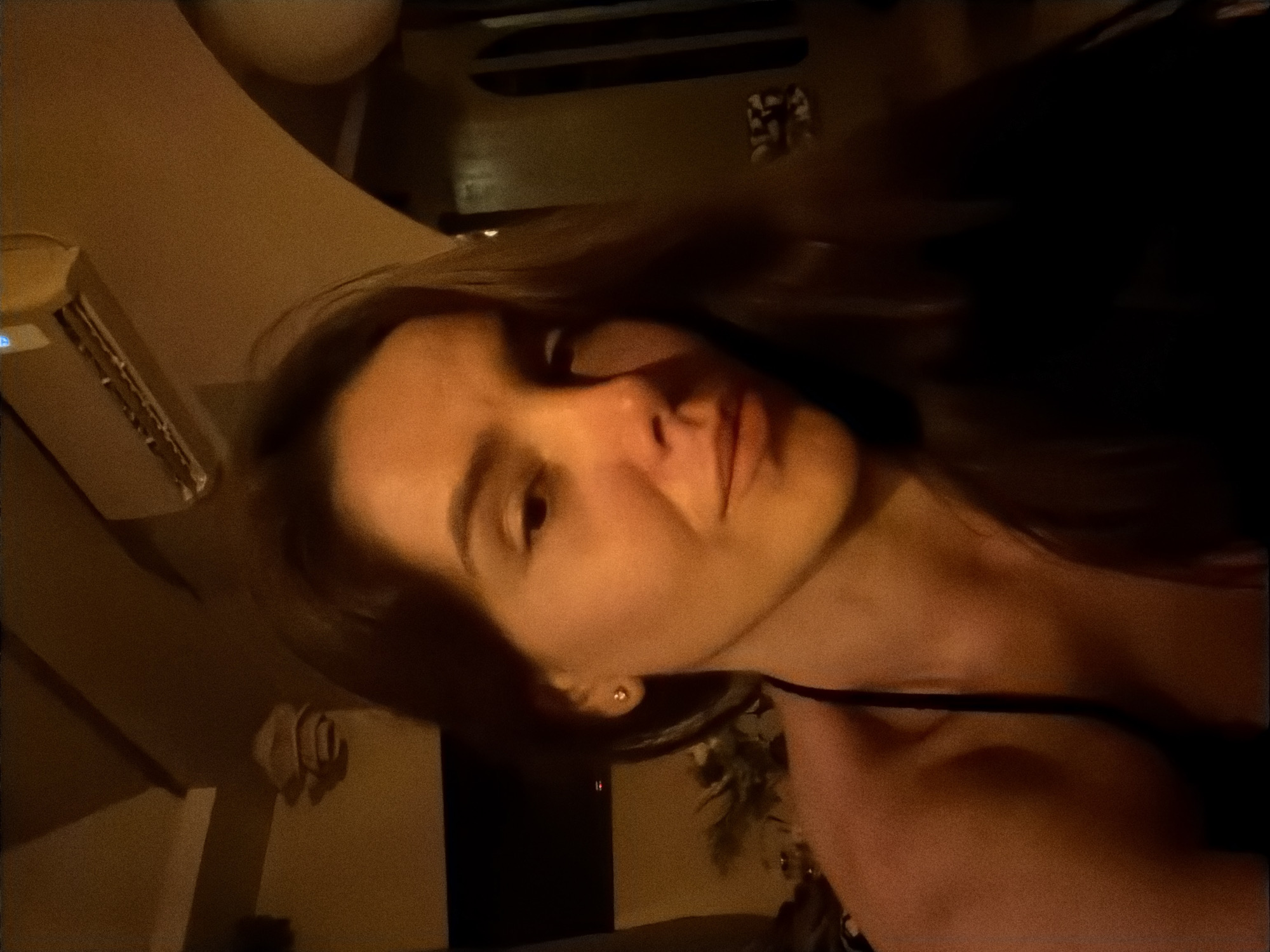 The image is a close-up, indoor, selfie-style photograph of a woman. She has long, dark brown hair, brown eyes, and light skin. She is wearing a black top. The background is dimly lit and appears to be a room with an air conditioning unit visible on the ceiling. There are also some indistinct objects in the background, including what looks like a decorative item on the wall and a dark, rectangular object that could be a television screen. The lighting is warm and appears to be artificial, creating a soft glow on the woman's face. The overall impression is one of a casual, personal moment captured in a domestic setting. The location in Mengwi, Indonesia, is not discernible from the visual information in the image.