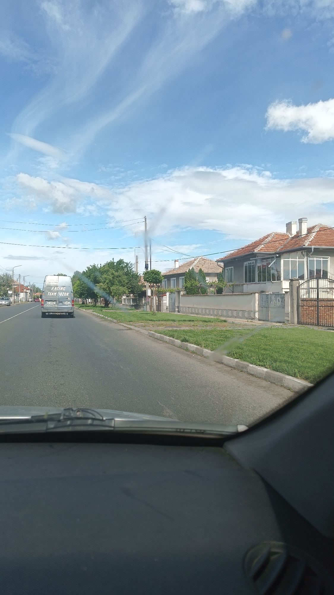 The image depicts a view from inside a vehicle, likely a car, traveling on a road in Popovitsa, Bulgaria. A grey van with the text "RACING TEAM OBZOR" on the back is visible ahead, moving in the same direction. The road is flanked by houses with tiled roofs and fenced yards with green grass. There are also trees lining the road. In the distance, a tall structure appears to be emitting dark smoke into the bright, blue sky with wispy clouds. The windshield of the vehicle obscures the bottom portion of the image.
