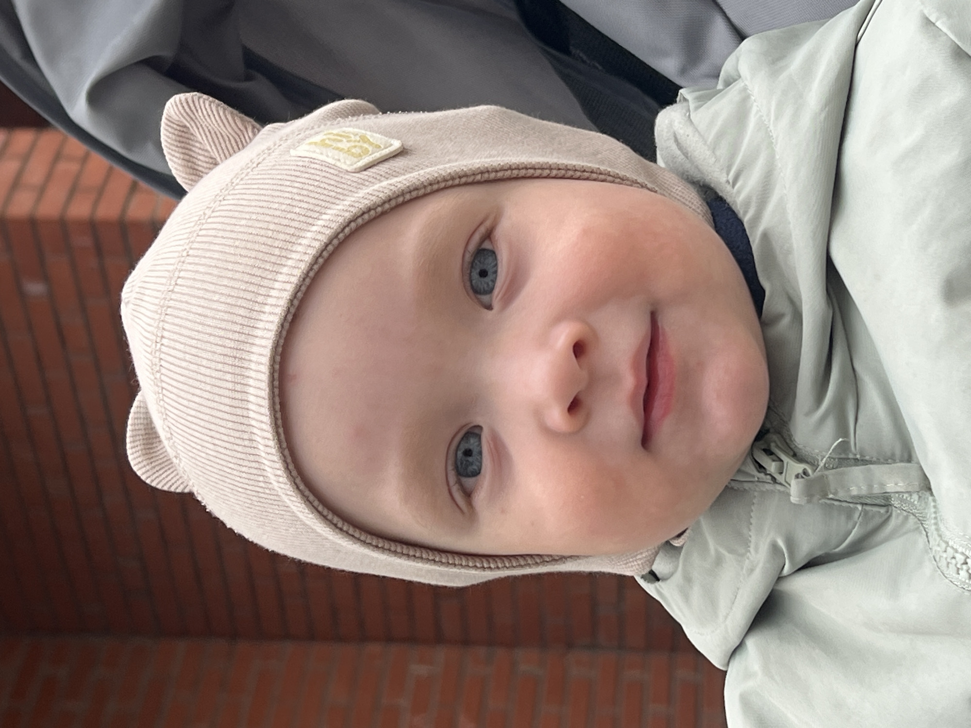 A close-up portrait features a young child, likely a baby or toddler, situated in the city of Chaikovsky, Russia. The child, with fair skin and blue eyes, looks directly at the camera with a slight smile. They are wearing a light beige, ribbed knitted bonnet with two prominent, rounded ear-like protrusions on top and a small, diamond-shaped yellow patch on the right side. Underneath the bonnet, a light green hooded jacket is visible, zipped partially. A dark blue collar is discernible beneath the jacket's hood. The child's cheeks exhibit a natural flush. The background consists of a red brick wall on the left side and the grey fabric of a stroller or carrier on the right.