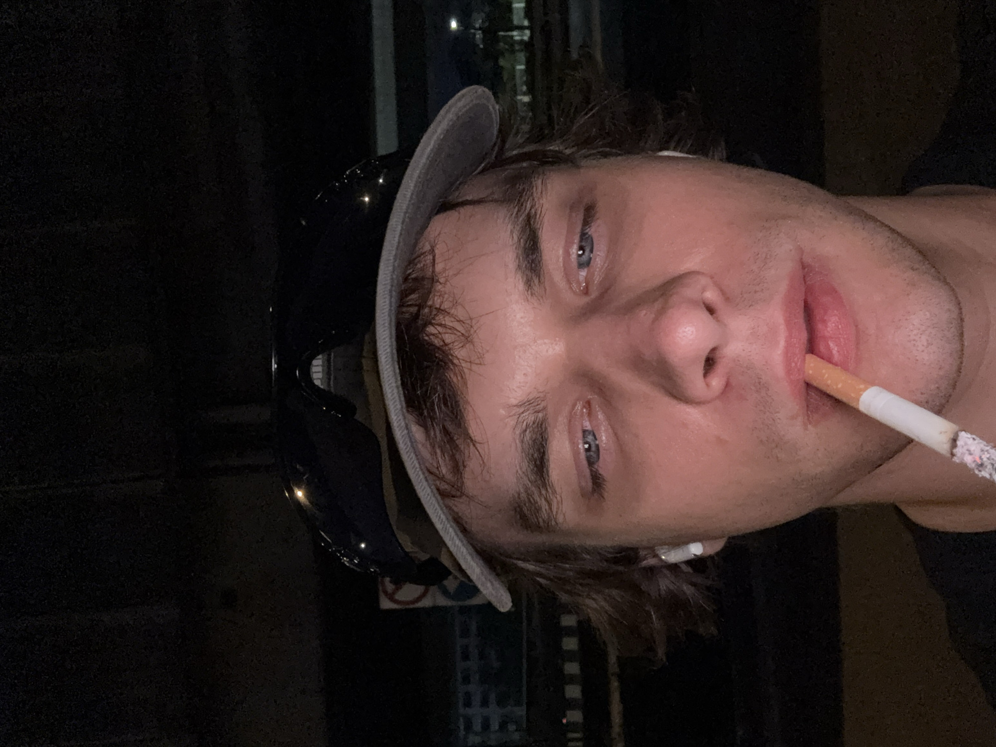 A close-up, low-light photograph shows a young, fair-skinned man holding a lit cigarette in his mouth. He is wearing a baseball cap and has what appears to be an earbud in his ear. The background suggests an urban setting, possibly at night, indicated by lights. Based on the photograph's location data, the image was taken in Bangkok, Thailand.