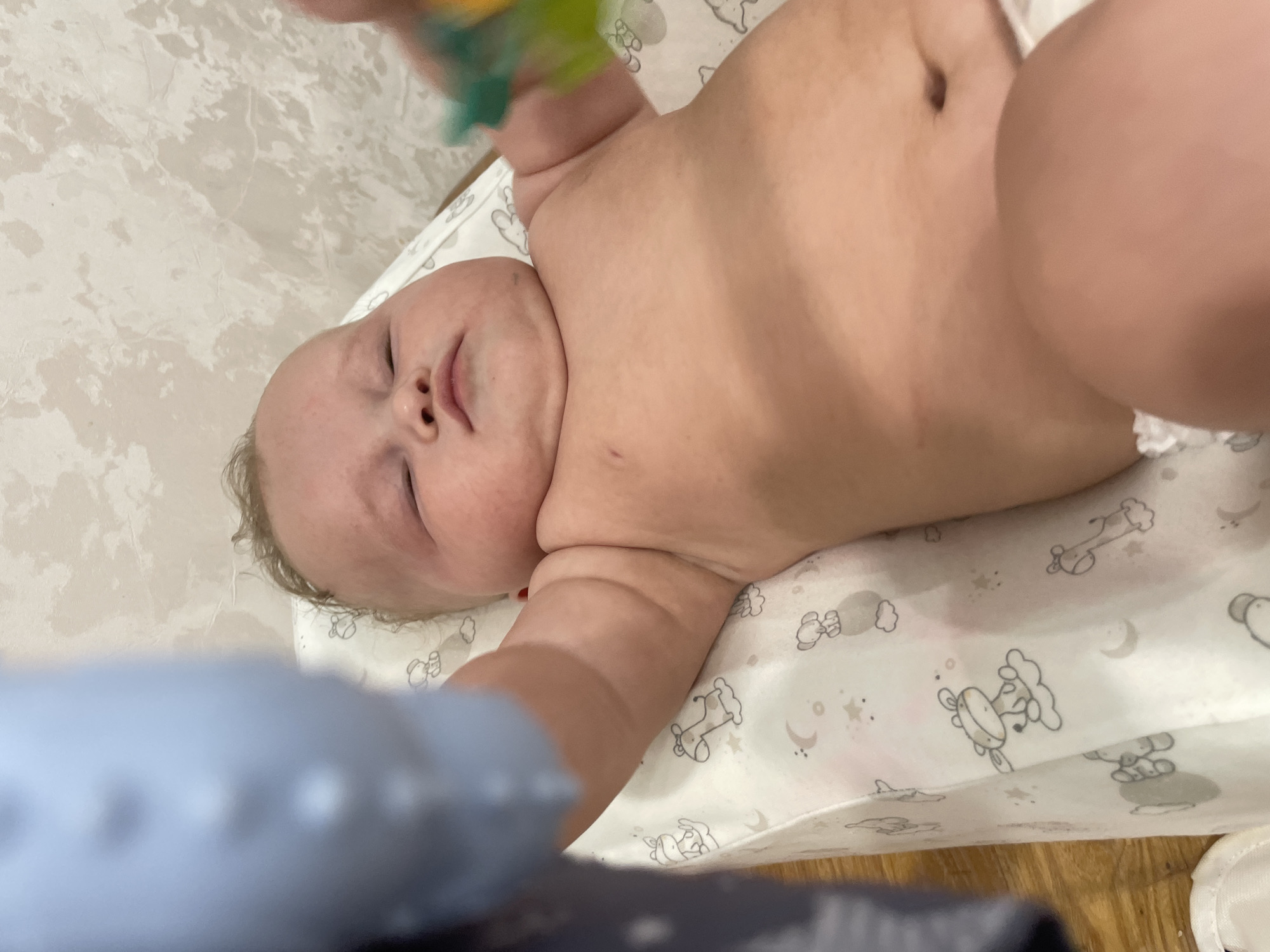 This is a close-up, overhead shot of a baby lying on its back. The baby's face is visible, with eyes closed and a slight smile. The baby's torso, arms, and legs are also in view, showcasing chubby rolls of skin. A colorful toy is partially visible at the top of the frame, held by an unseen hand. The baby is lying on a changing pad with a pattern of cartoon animals, clouds, and stars. The background shows a marbled surface and a wooden floor. The scene appears to be indoors, possibly a nursery or changing area. The lighting is soft, suggesting it's daytime. The overall mood is peaceful and intimate. No visible text is present in the image.