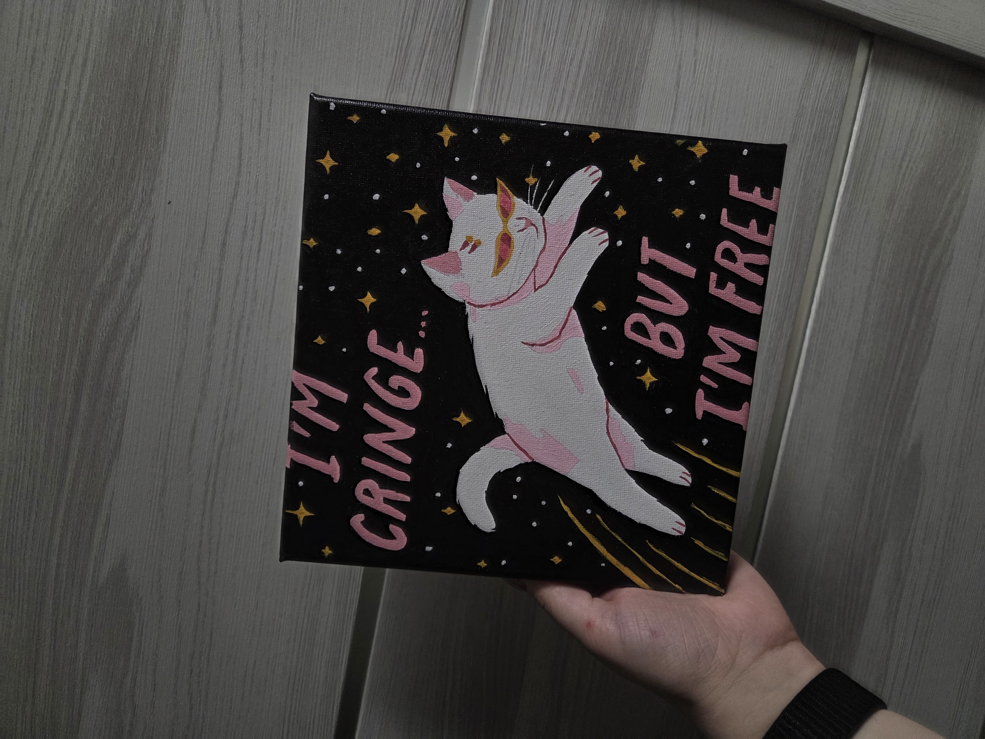 A hand holds a square black canvas with a painting of a white cat with pink accents wearing sunglasses. The cat is mid-leap, positioned diagonally on the canvas. Around the cat are yellow stars and white dots against the black background. In pink lettering, the words "I'M CRINGE..." are written on the left and "...BUT I'M FREE" on the right. Yellow lines radiate from the lower left corner of the canvas. The hand holding the canvas has a black wristband. The canvas is held in front of a gray wood-paneled wall. Location: Unknown, Unknown.